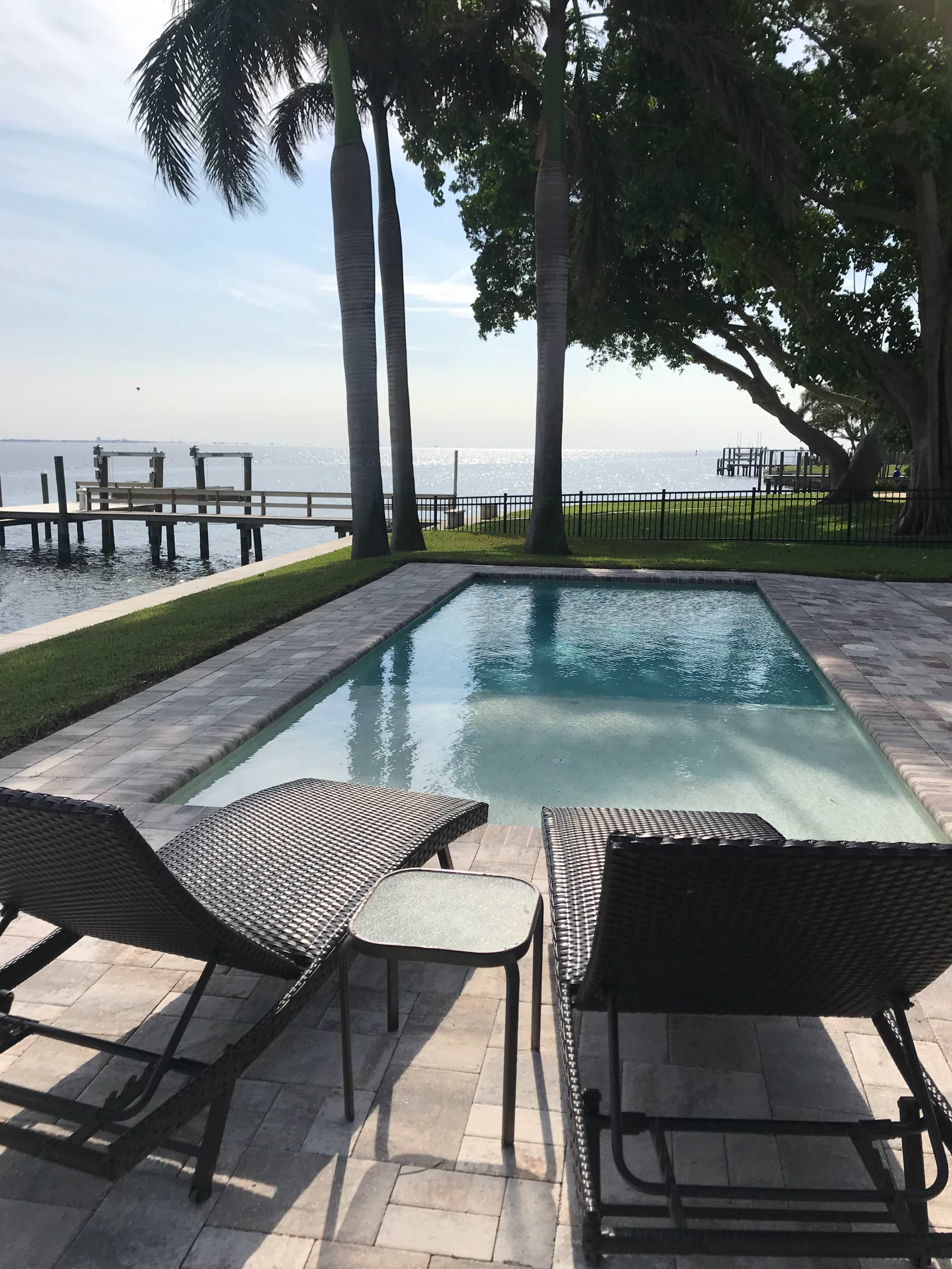 A poolside area features two lounge chairs next to a clear pool, with palm trees and a view of the water in the background.