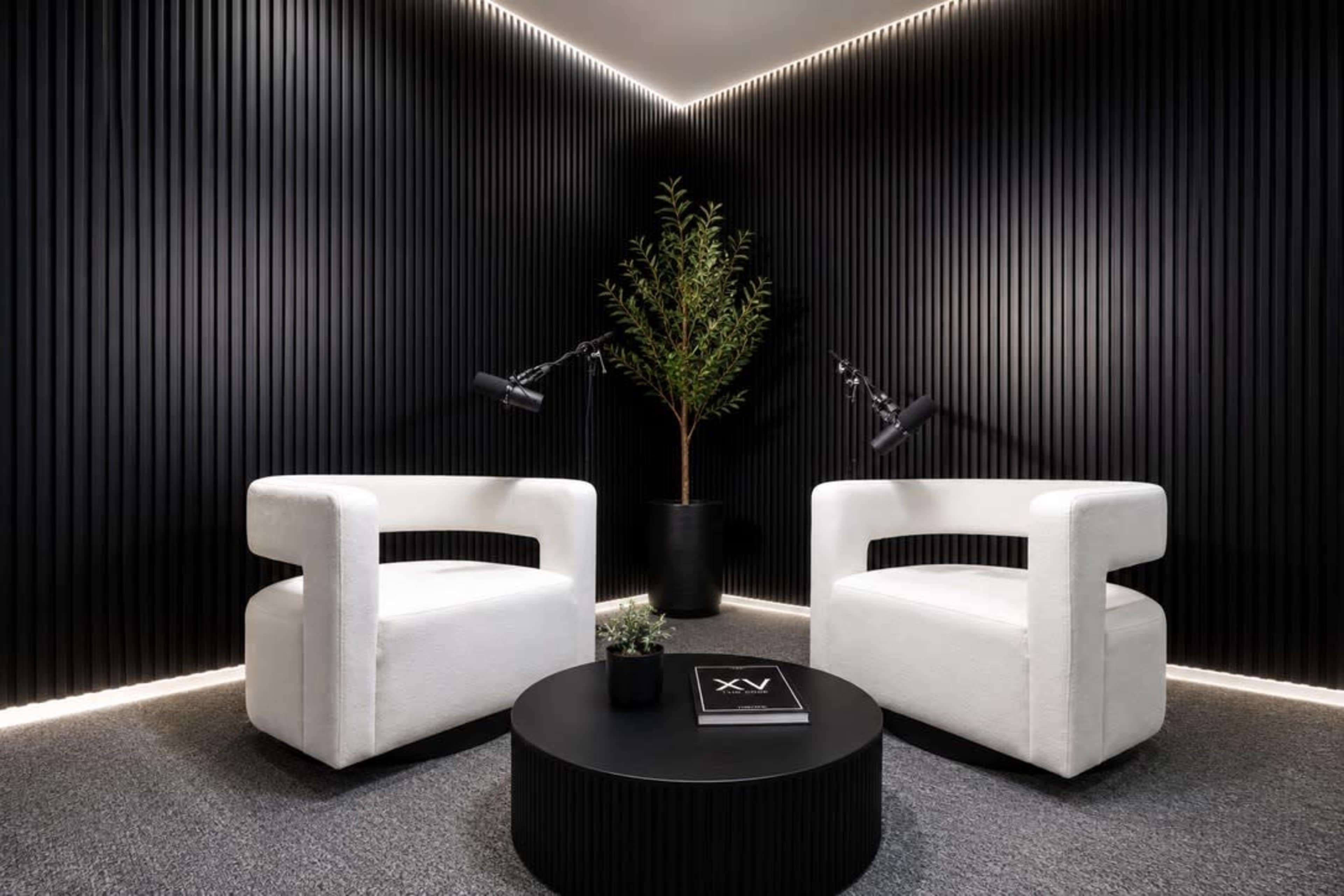The image features two white, modern chairs positioned across from each other in a sleek, black-paneled room with a round black coffee table and a potted plant in the corner.