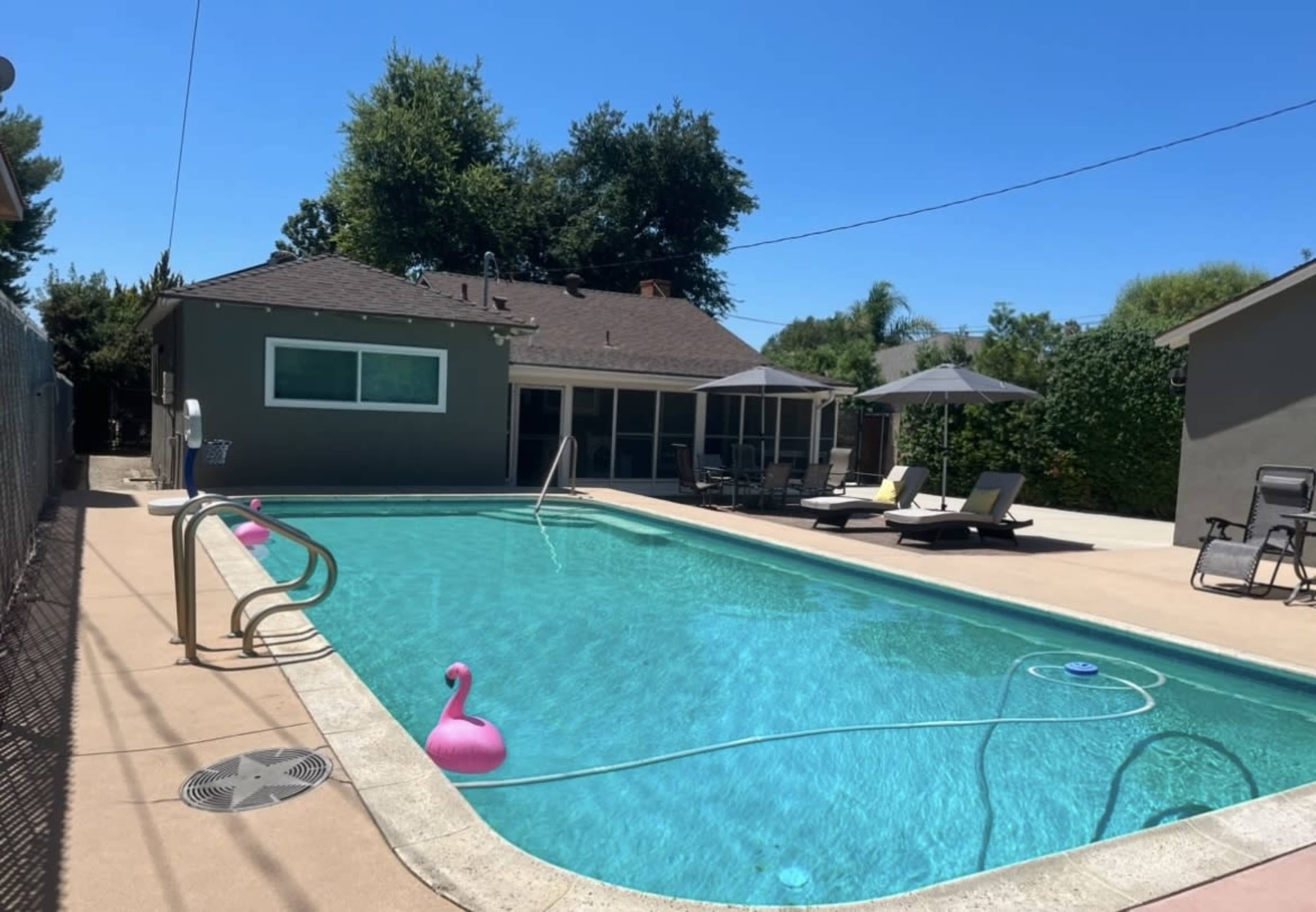 A clear blue swimming pool is situated behind a house, surrounded by lounge chairs and umbrellas, with a fence enclosing the area.