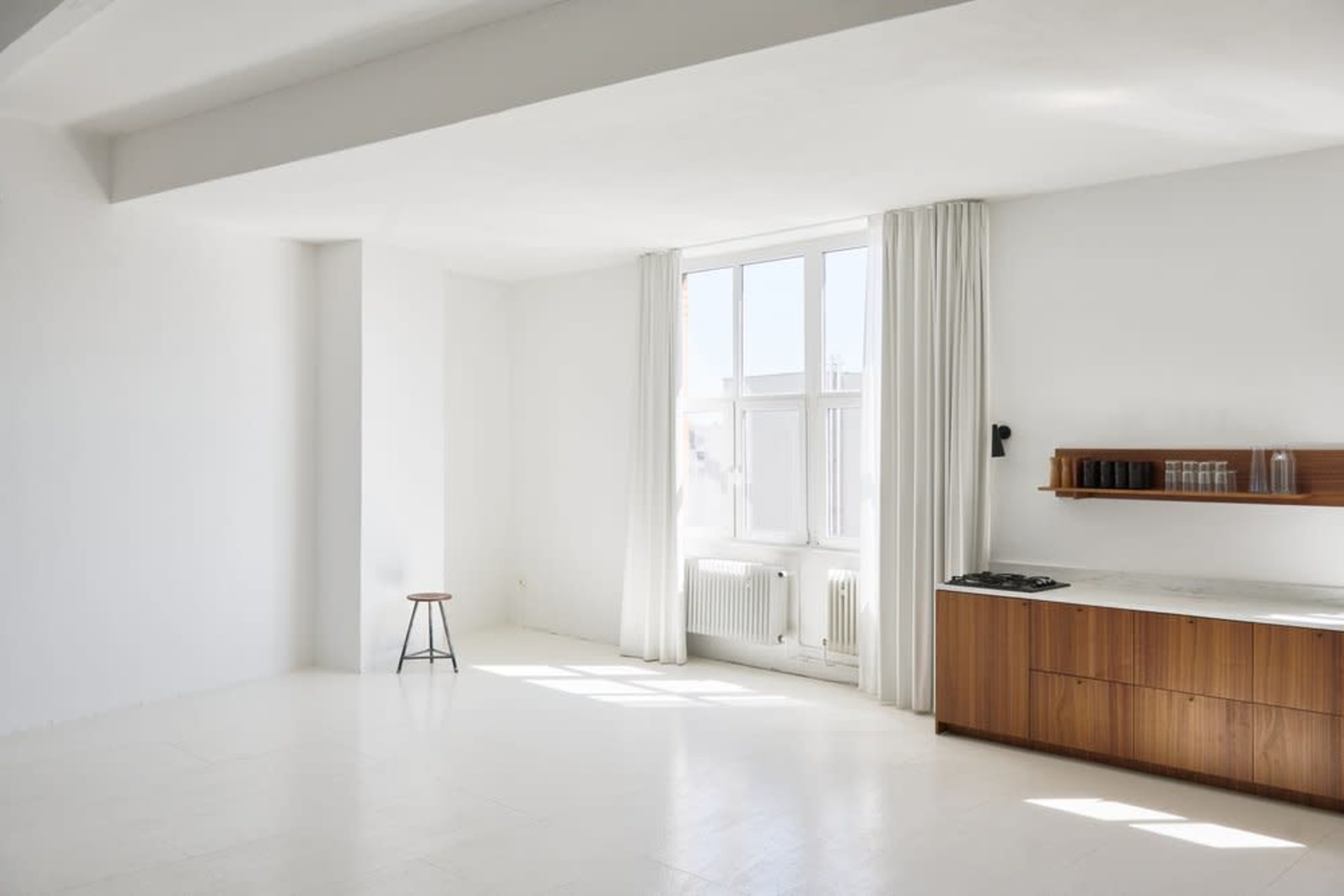 The image depicts a spacious, minimalistic room featuring a large window, white walls, a wooden countertop, and a stool.