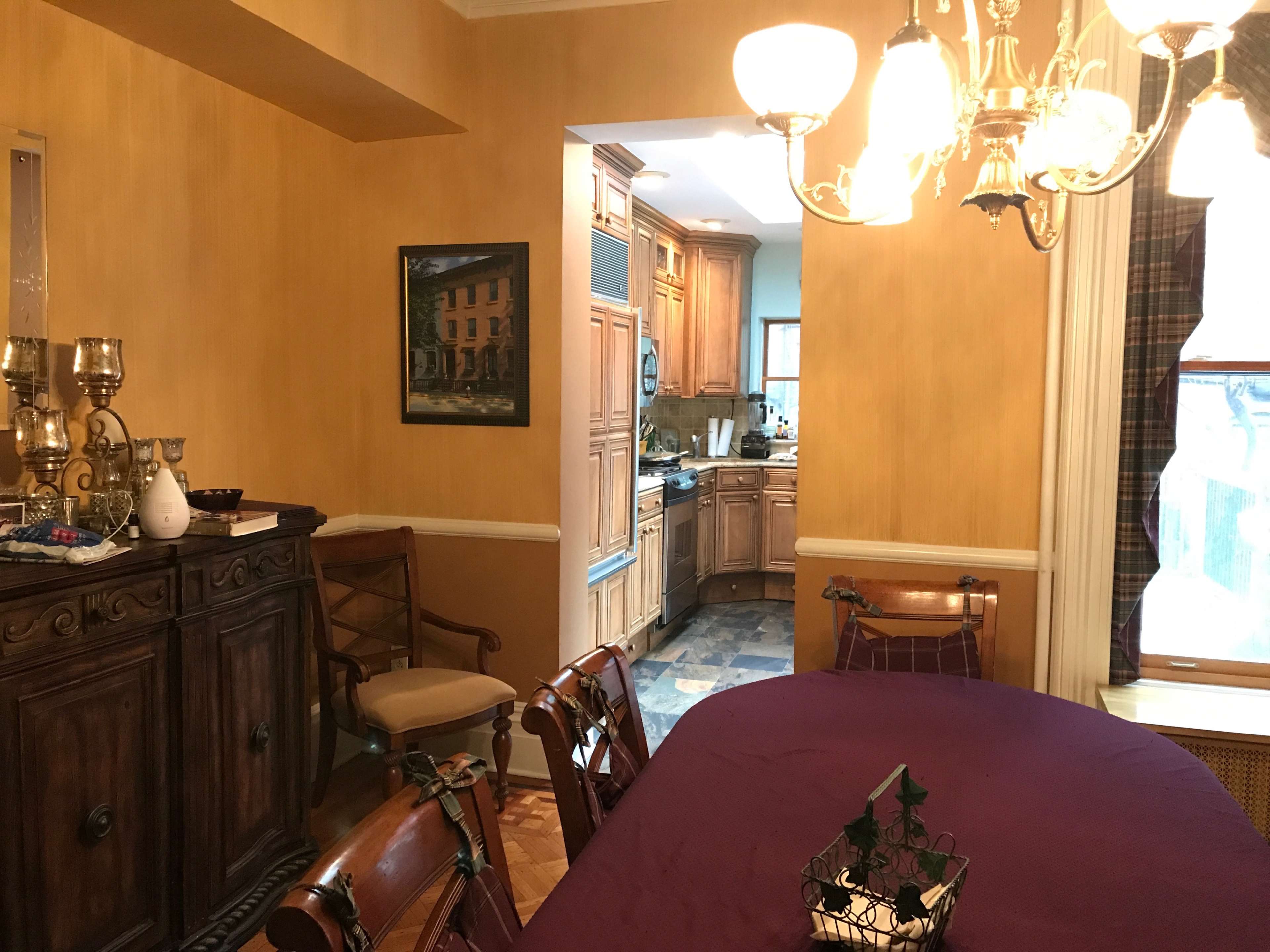 A dining room with a wooden table and chairs, adorned with a purple tablecloth, and a view into a kitchen through an open doorway.