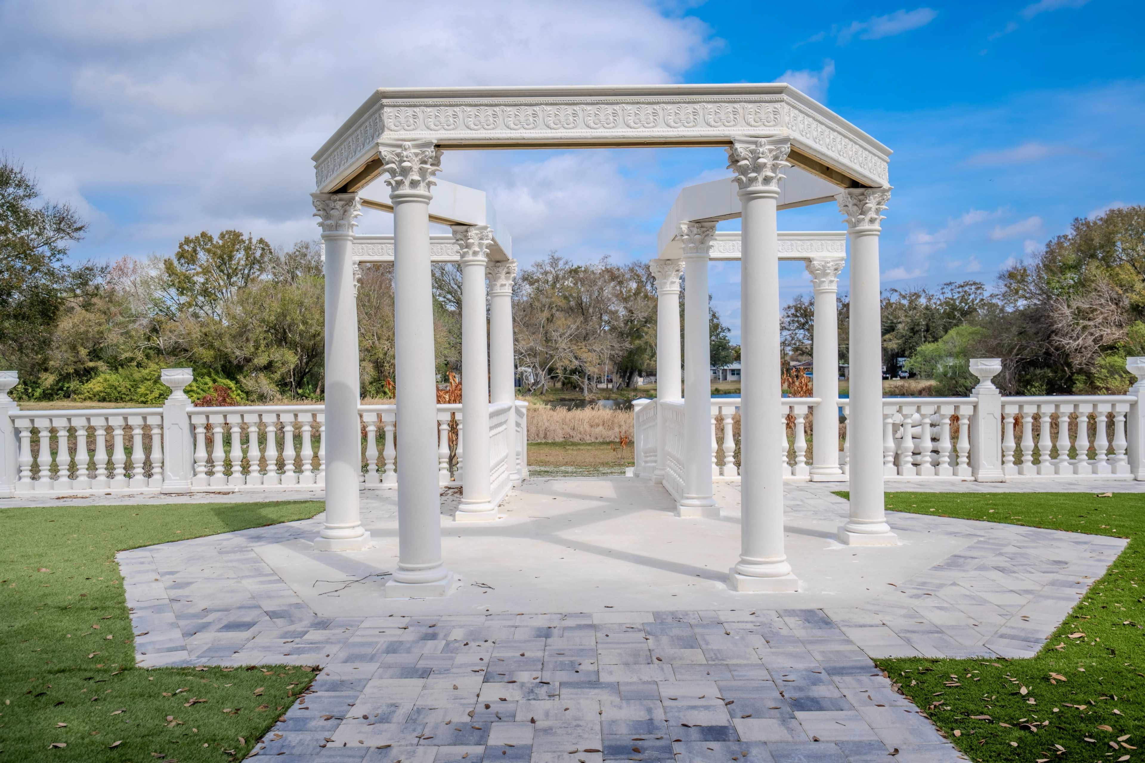 The image shows a white, columned gazebo situated on a stone platform surrounded by a grassy area and trees in the background.