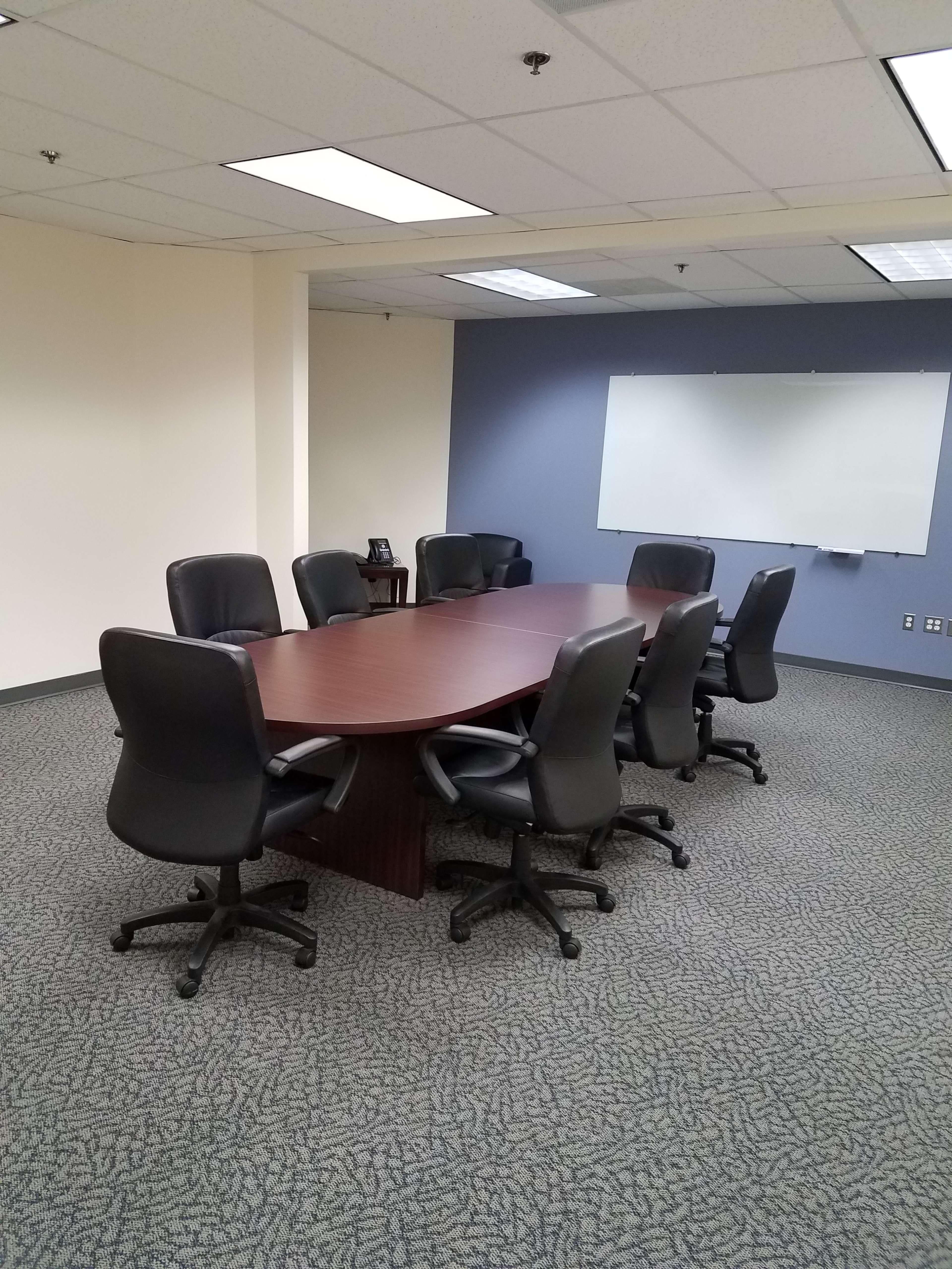 A long rectangular conference table is surrounded by eight black office chairs in a sparsely furnished meeting room with a whiteboard on one wall.
