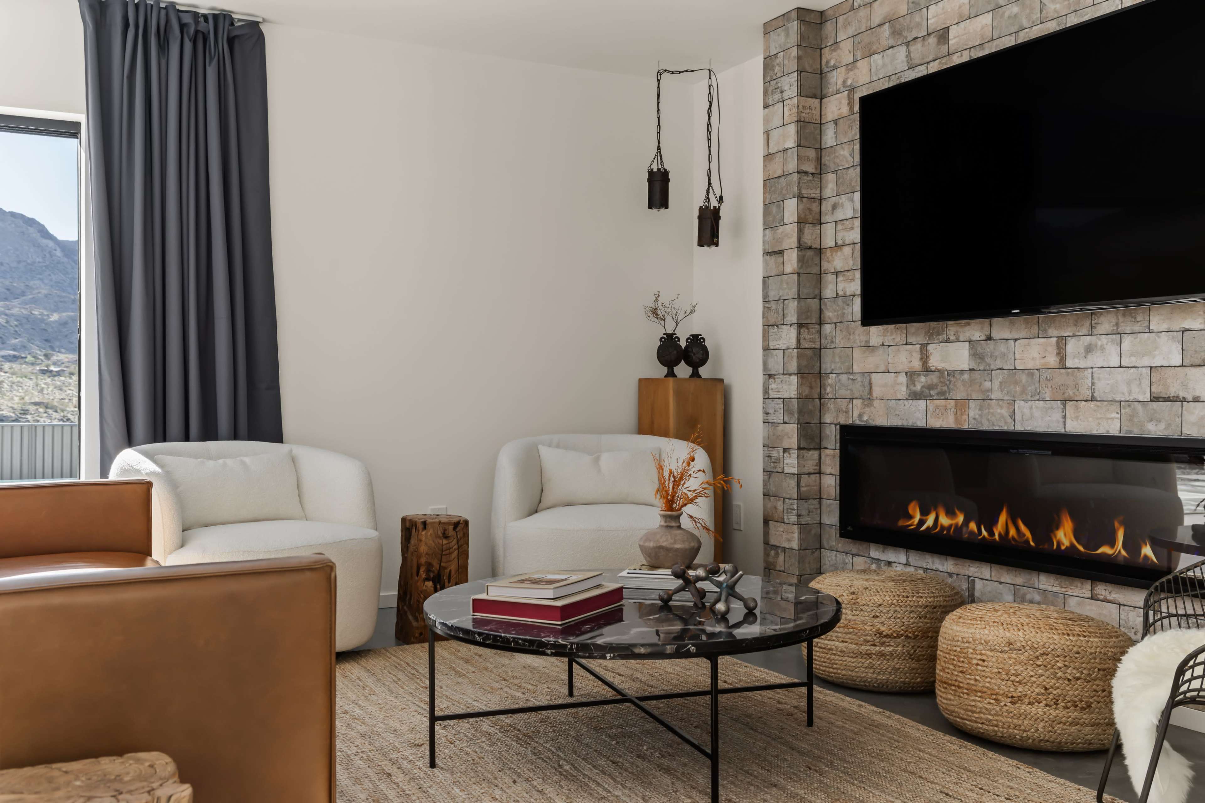 The image shows a modern living room with two white armchairs, a round black coffee table, a television mounted on a stone accent wall, and a fireplace beneath it.