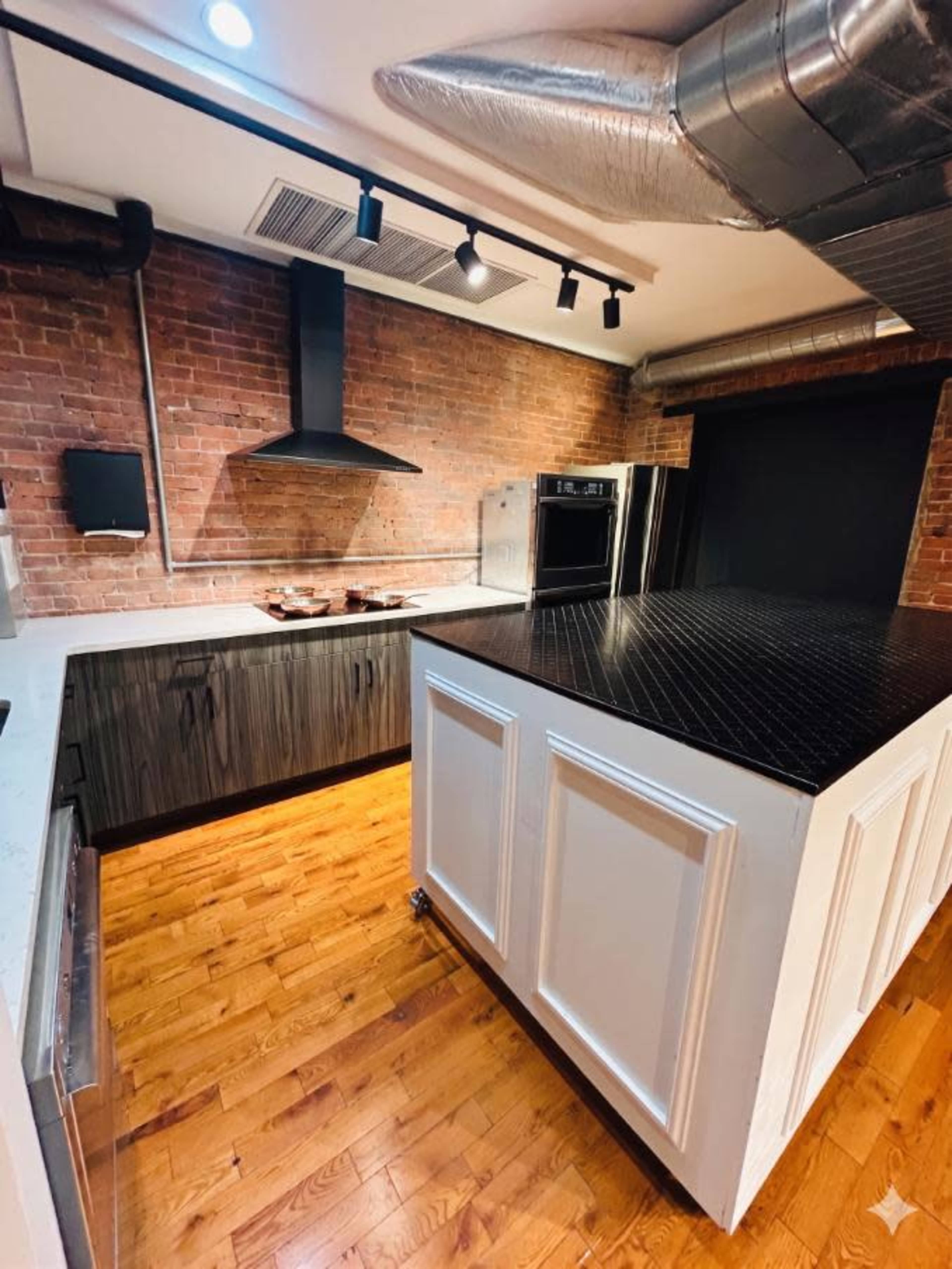 The image shows a modern kitchen with exposed brick walls, a dark cabinetry layout, a large black countertop island, and stainless steel appliances.
