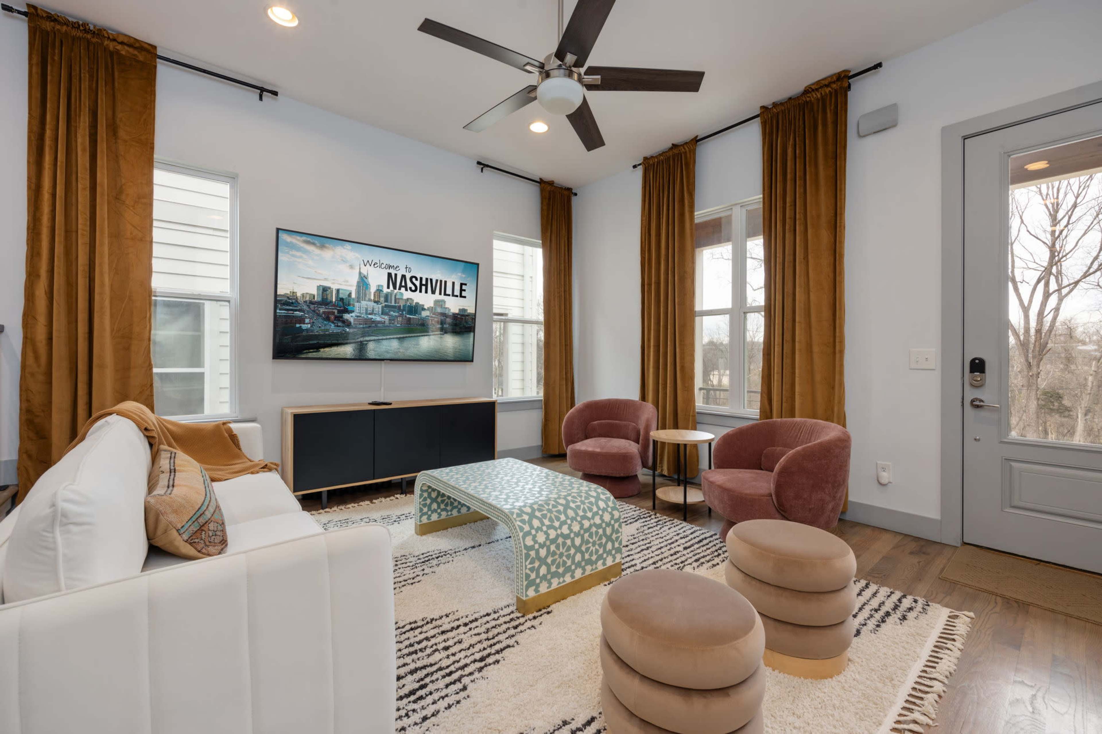 The living room features a television mounted on the wall, framed by large windows and decorated with elegant curtains, a white sofa, pink armchairs, and a patterned coffee table.