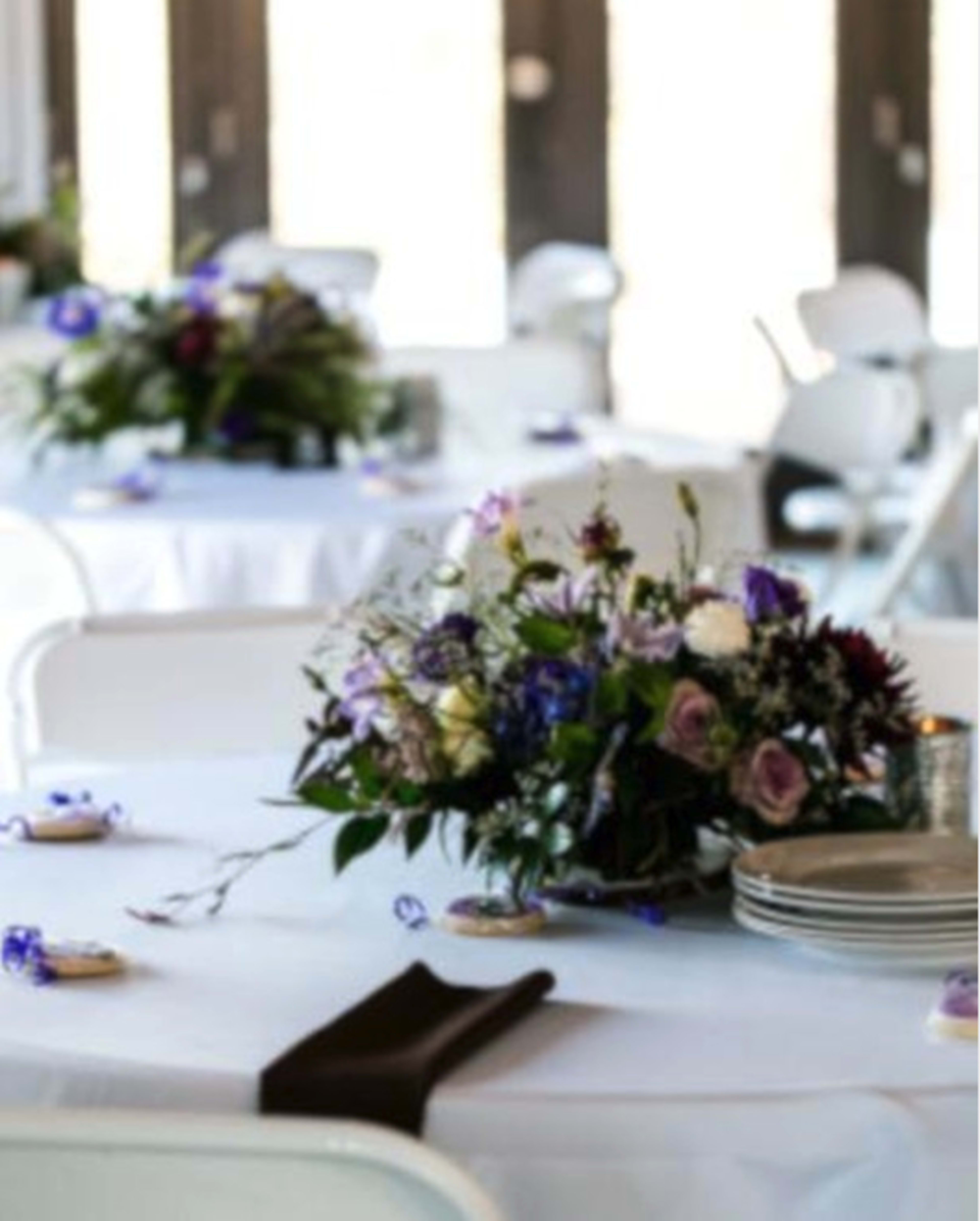 A white-tableclothed dining setup features several round tables adorned with floral centerpieces in shades of purple and green.
