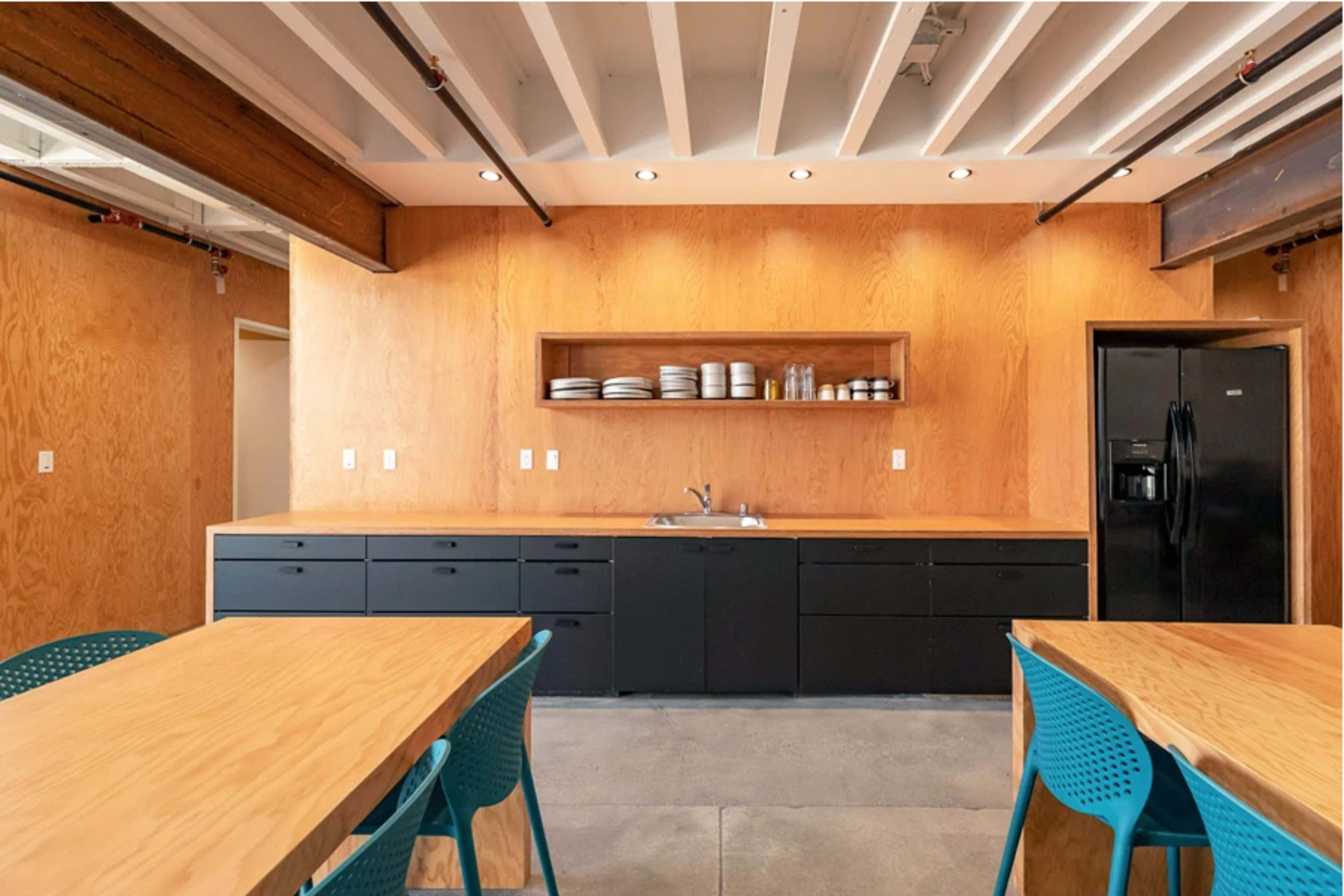 A modern kitchen with wooden walls, black cabinets, a sink, and shelves displaying plates and glassware, accompanied by two wooden tables and blue chairs.