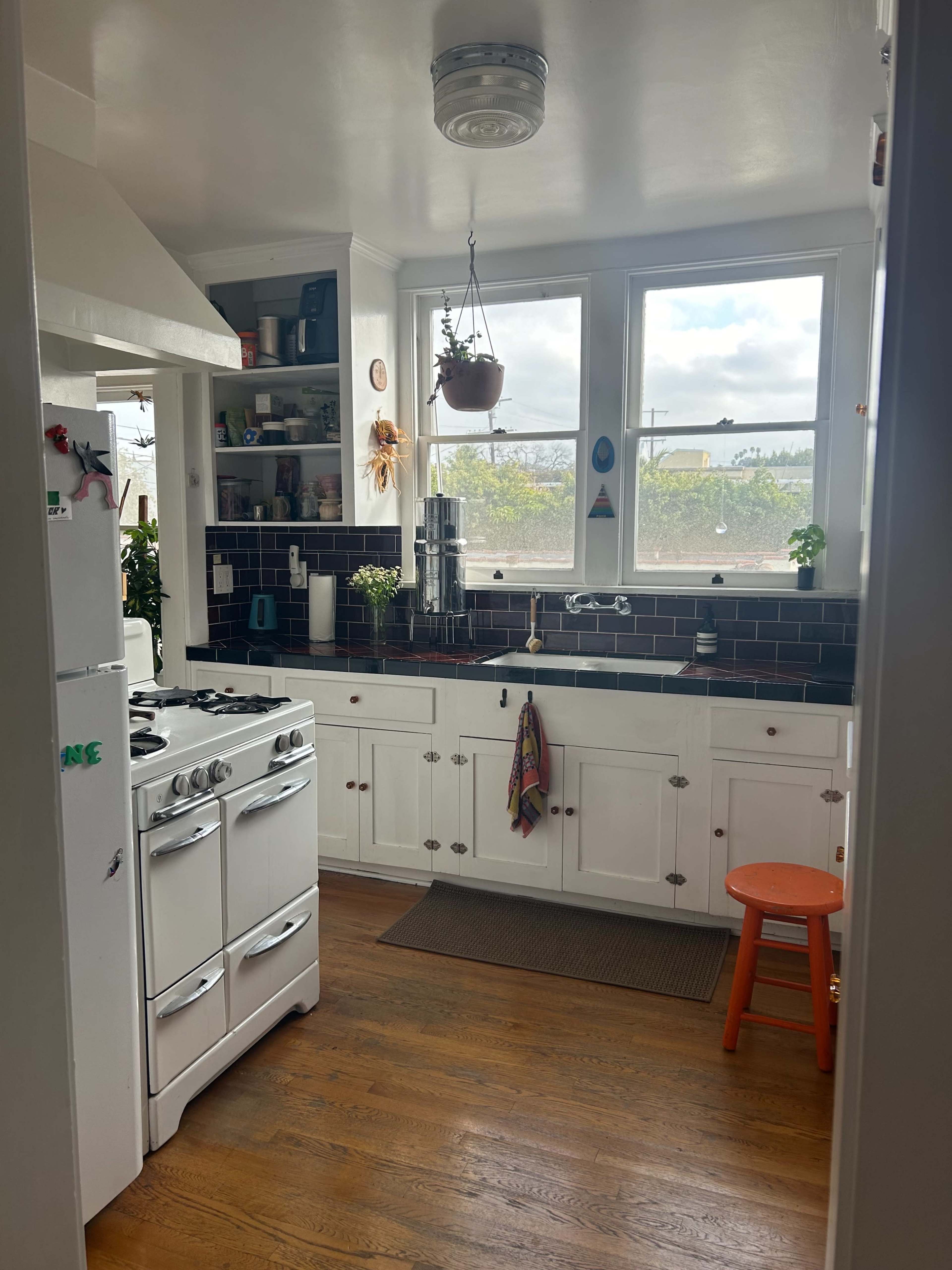 A bright kitchen features white cabinetry, a vintage stove, and large windows with plants on the sill, along with a wooden floor and an orange stool.