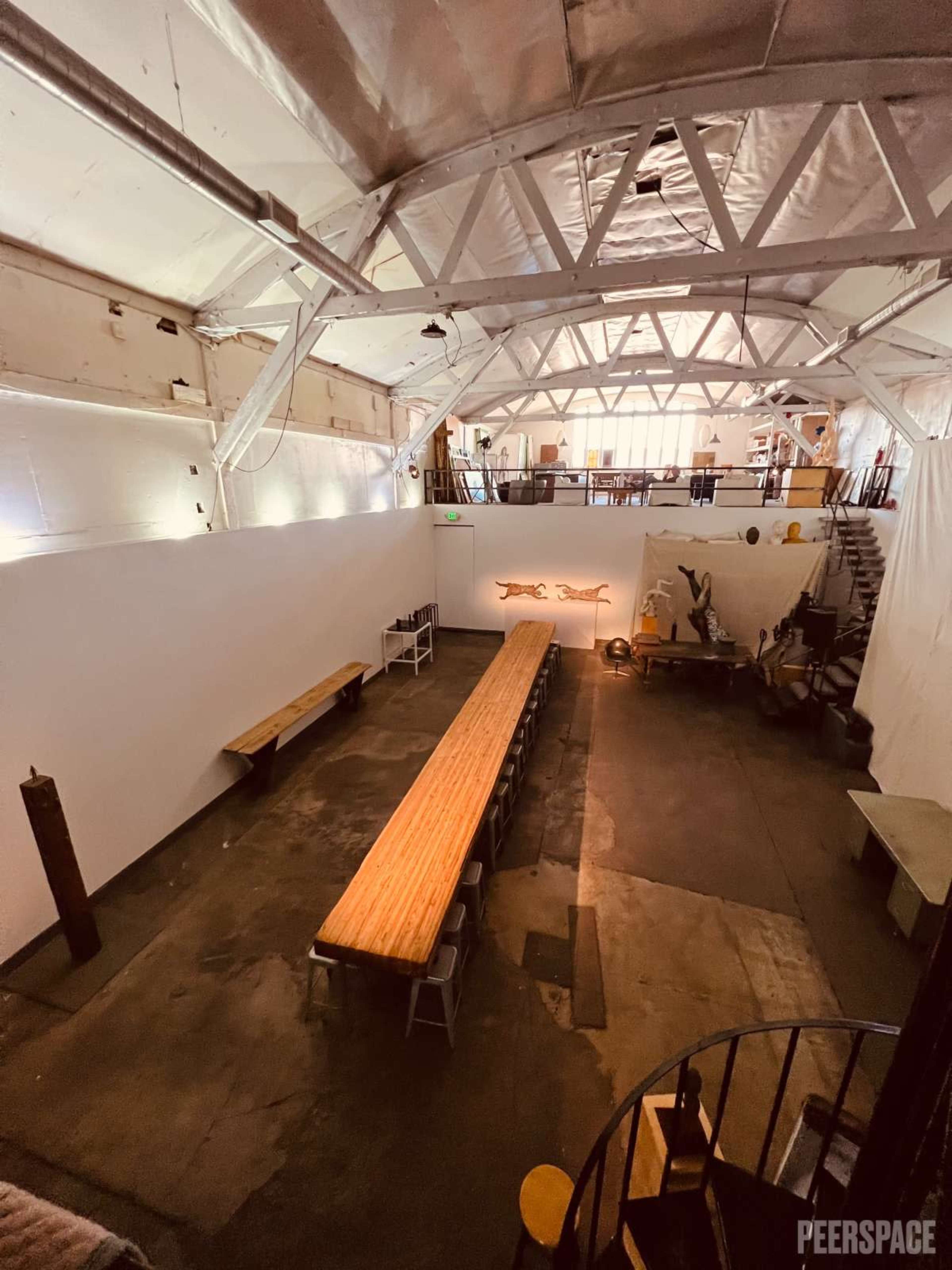 The image shows a spacious indoor venue with a long wooden table set for dining, surrounded by white walls and high, exposed beams.