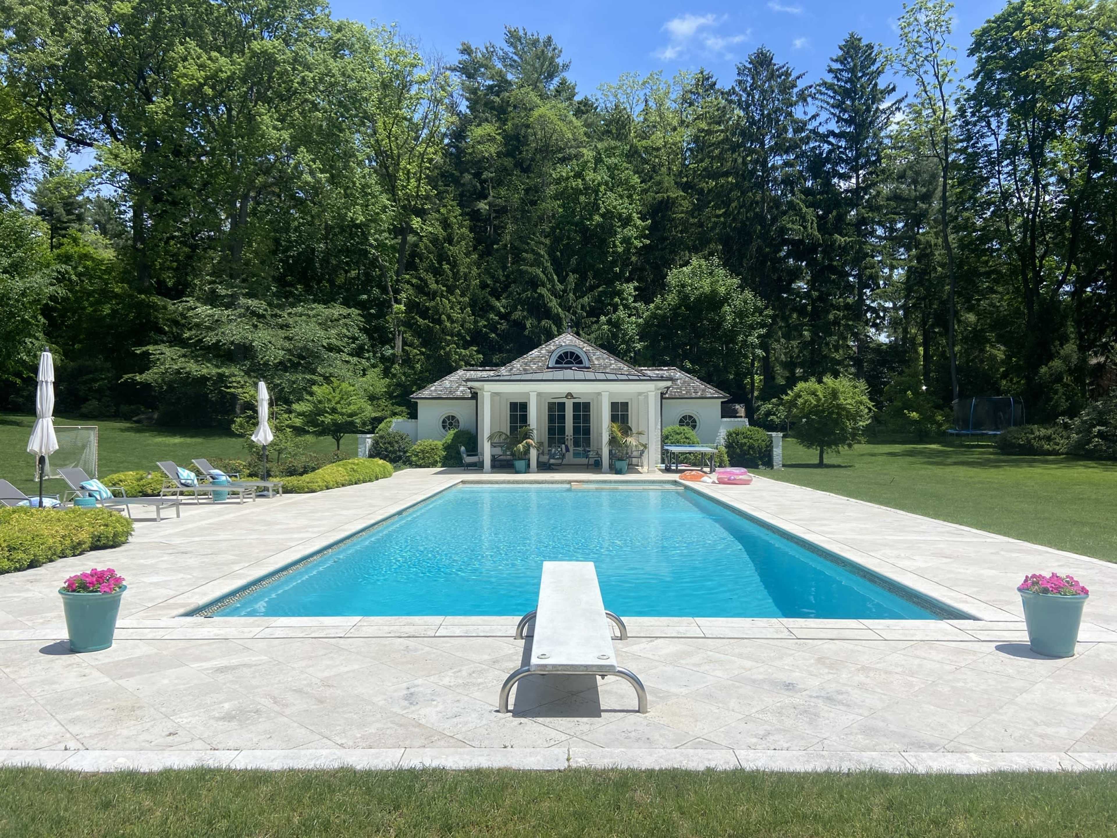A rectangular swimming pool is surrounded by a manicured lawn, lounge chairs, and potted plants, with a white gazebo visible in the background against a backdrop of trees.