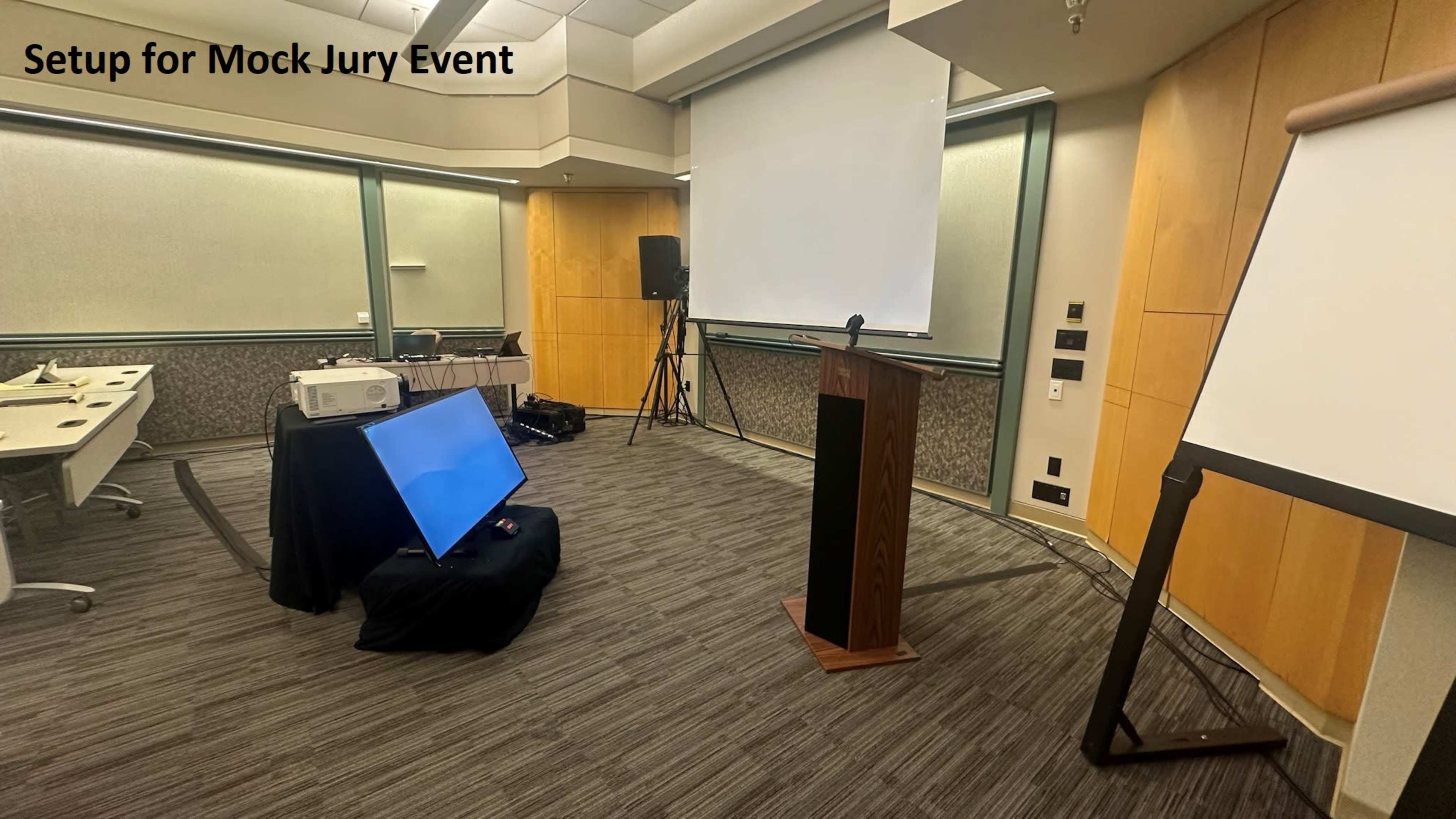 The image shows a conference room set up for a mock jury event, featuring a podium, projector, screen, and seating arrangement.