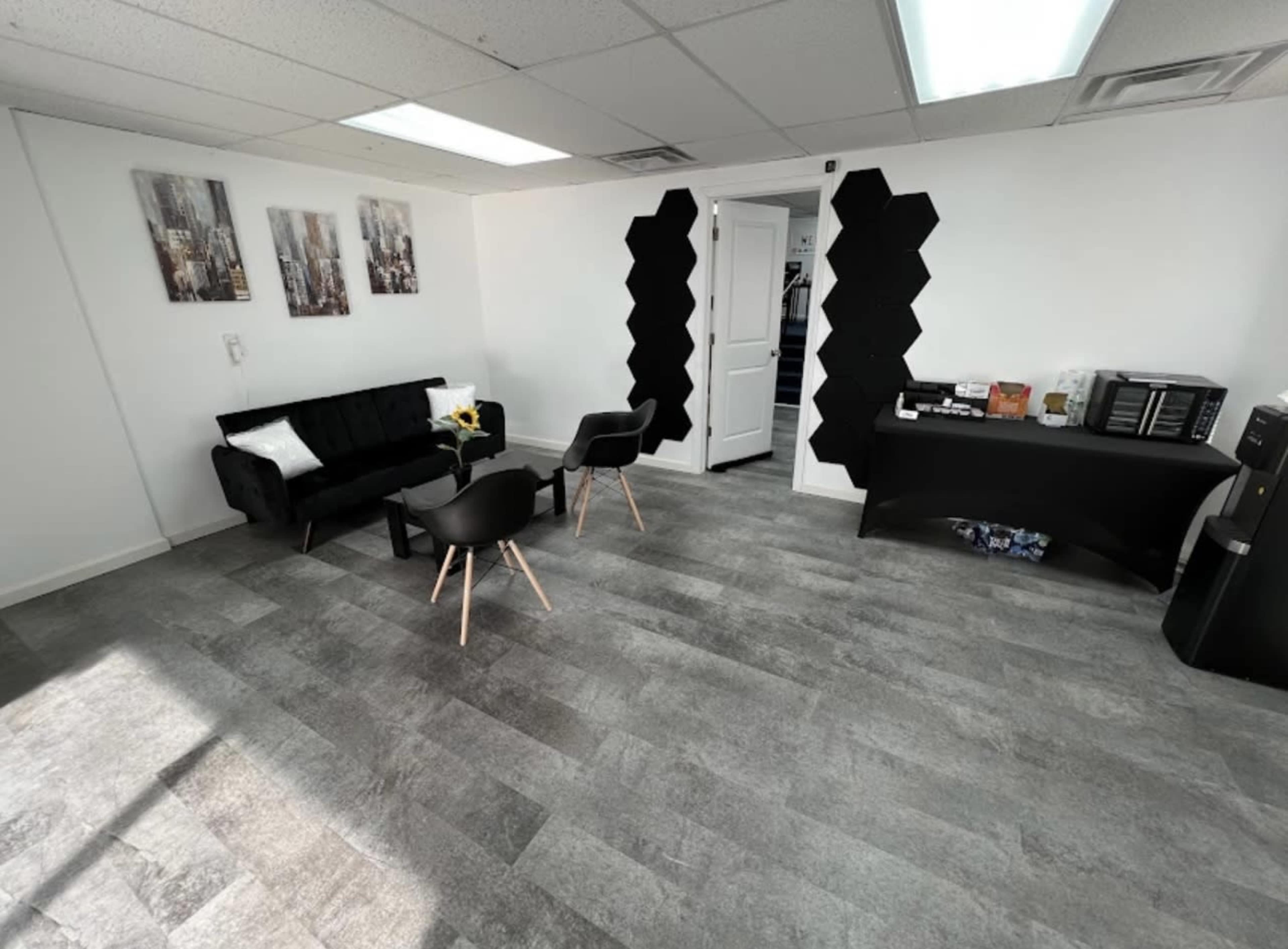 The room features a black couch, two black chairs, a side table, and a dark reception desk against a light-colored wall with abstract wall art.