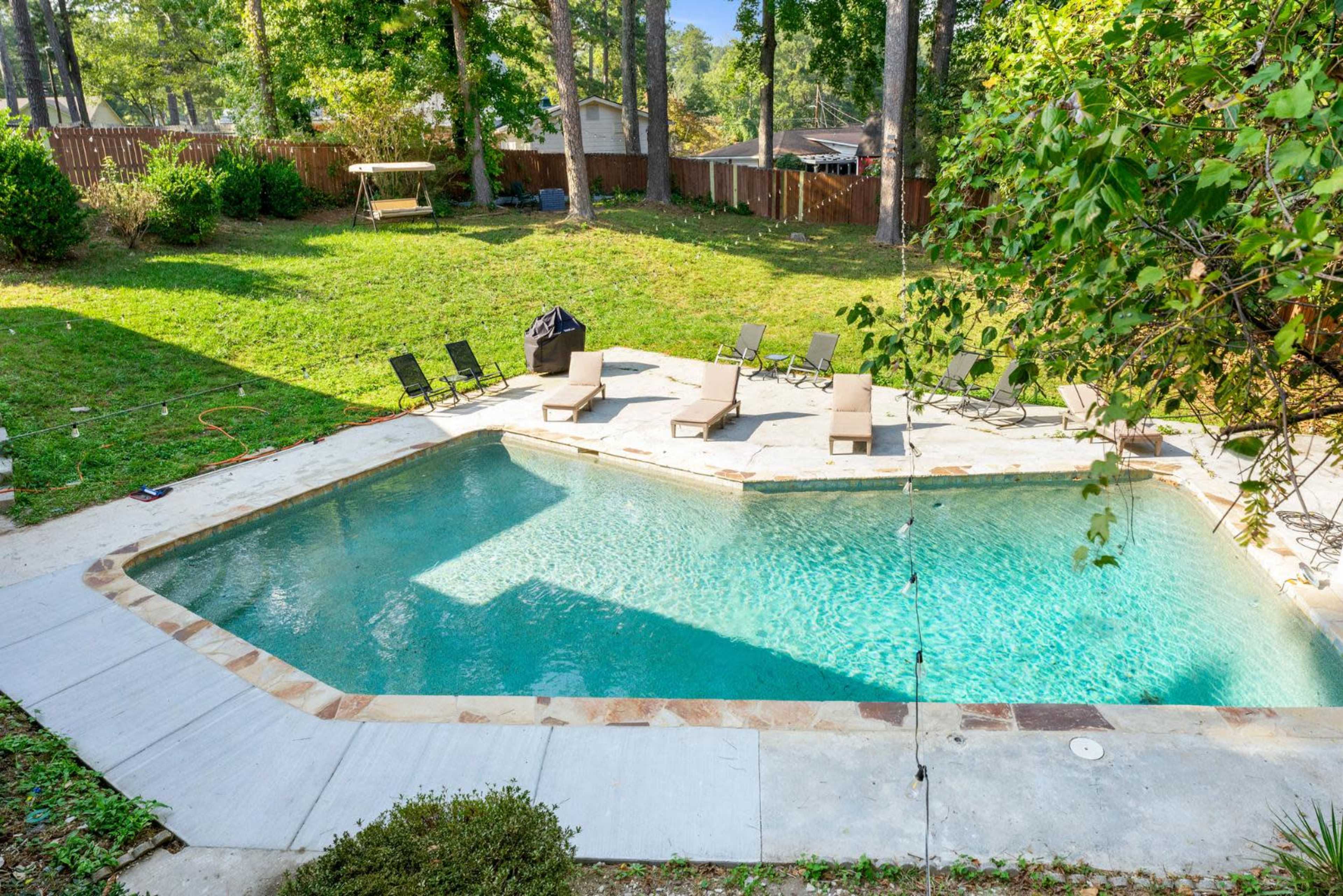 The image shows a backyard swimming pool surrounded by lounge chairs and a grassy area with trees and a swing set in the background.