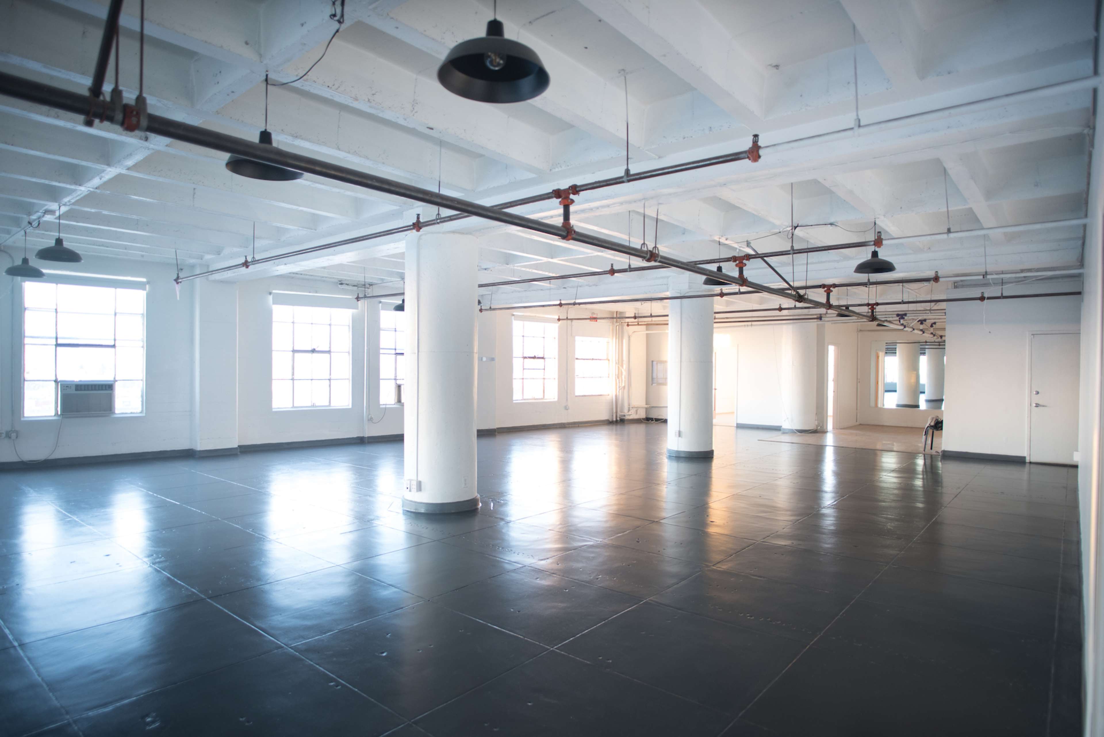 A spacious, empty industrial-style room with polished concrete floors and large windows, illuminated by overhead lights.