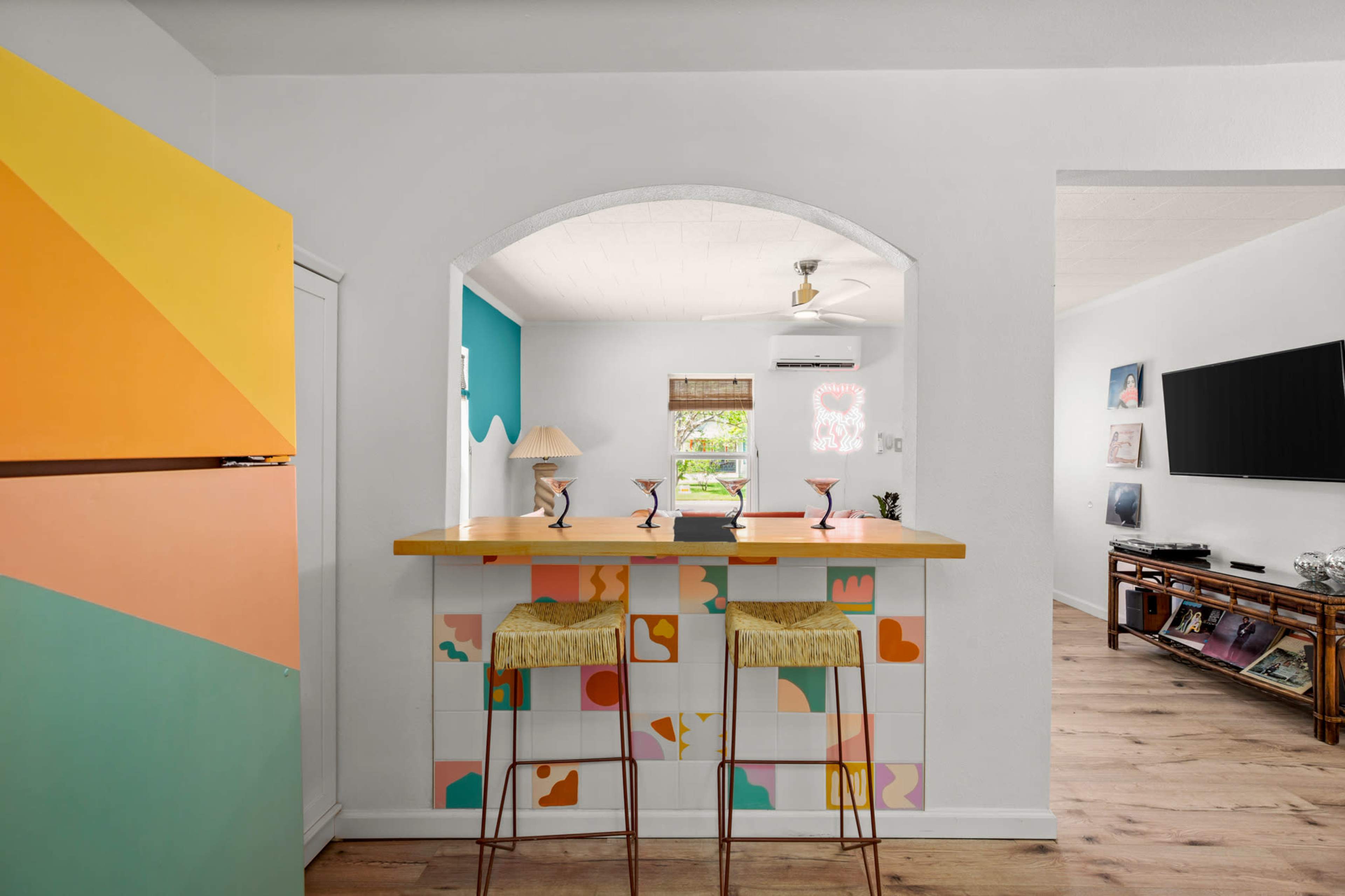 A colorful kitchen features a bright refrigerator, a bar with two stools, and an open layout leading to a living area.
