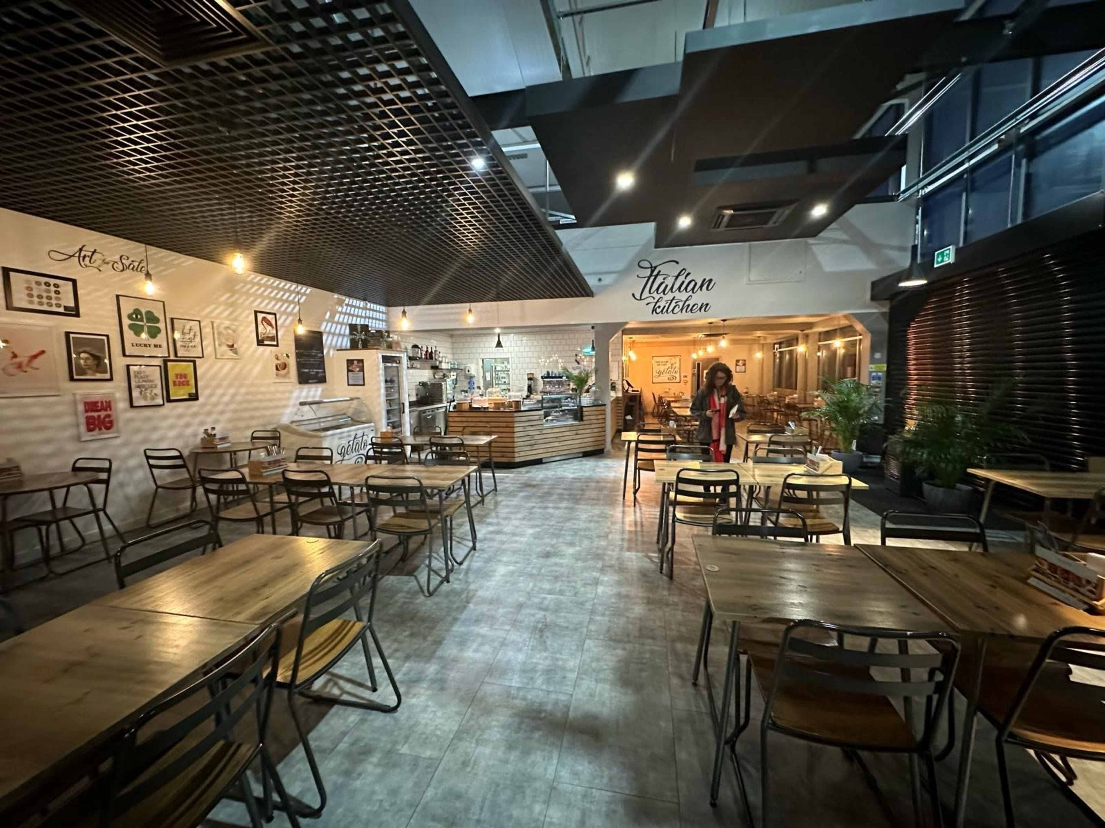 A spacious, indoor restaurant with wooden tables and chairs, artwork on the walls, and a visible kitchen area at the back.