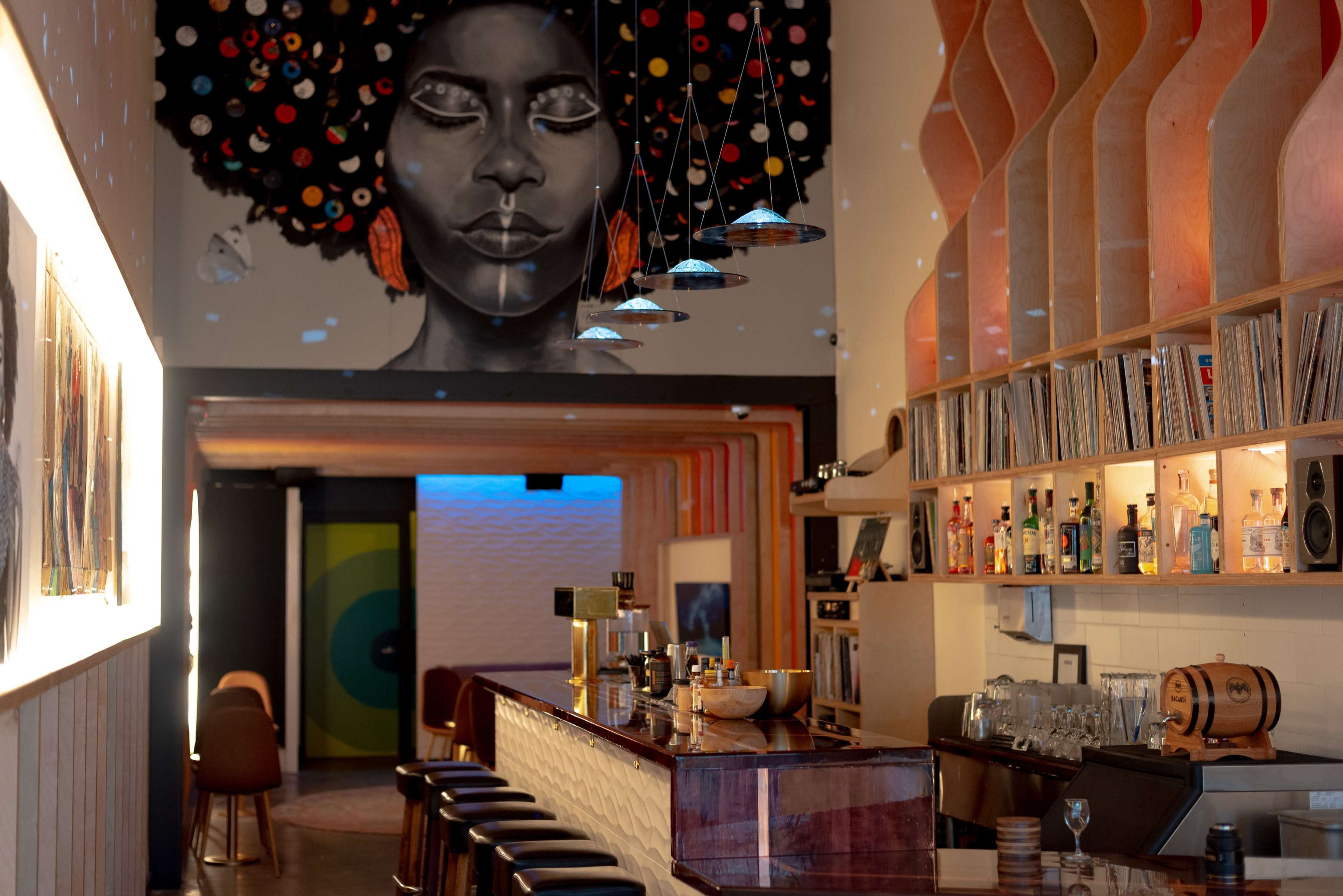 A spacious bar with a vibrant mural of a woman's face on the wall, surrounded by warm wooden accents and a well-stocked shelf of beverages.