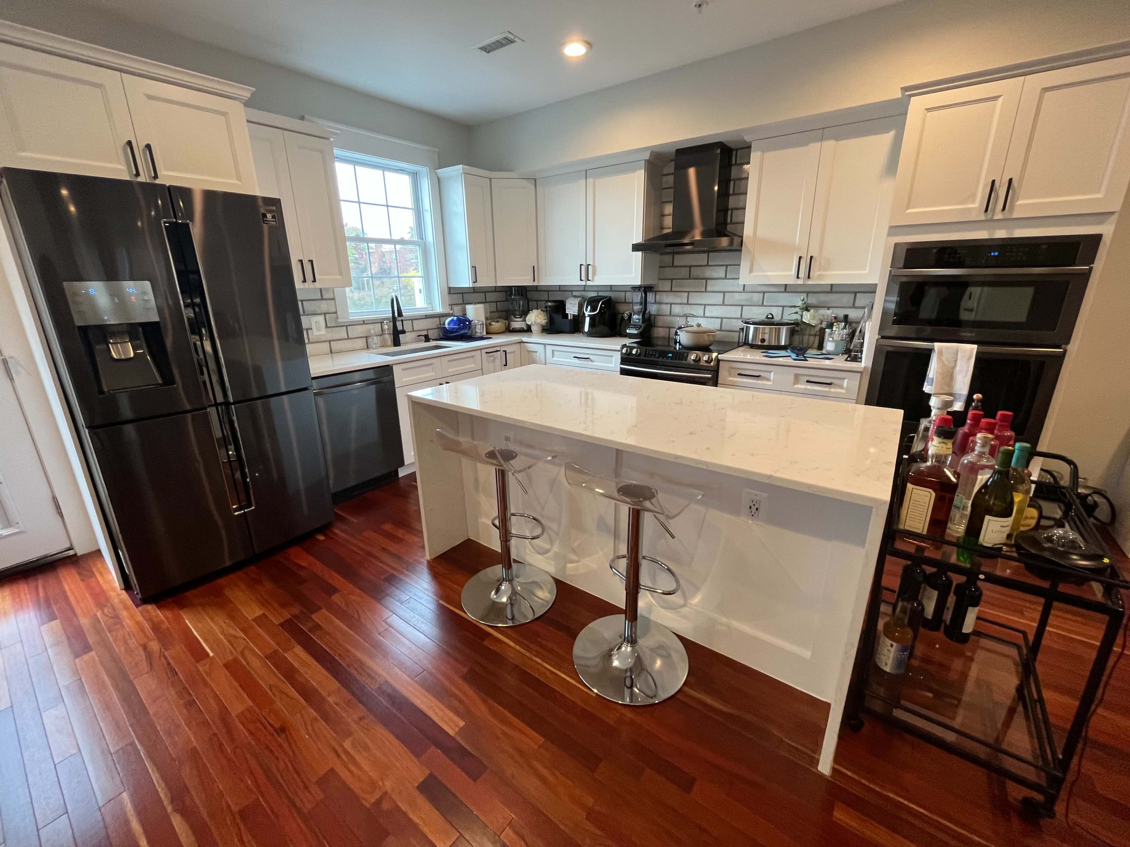 The image shows a modern kitchen with white cabinets, stainless steel appliances, and a central island equipped with two clear bar stools.