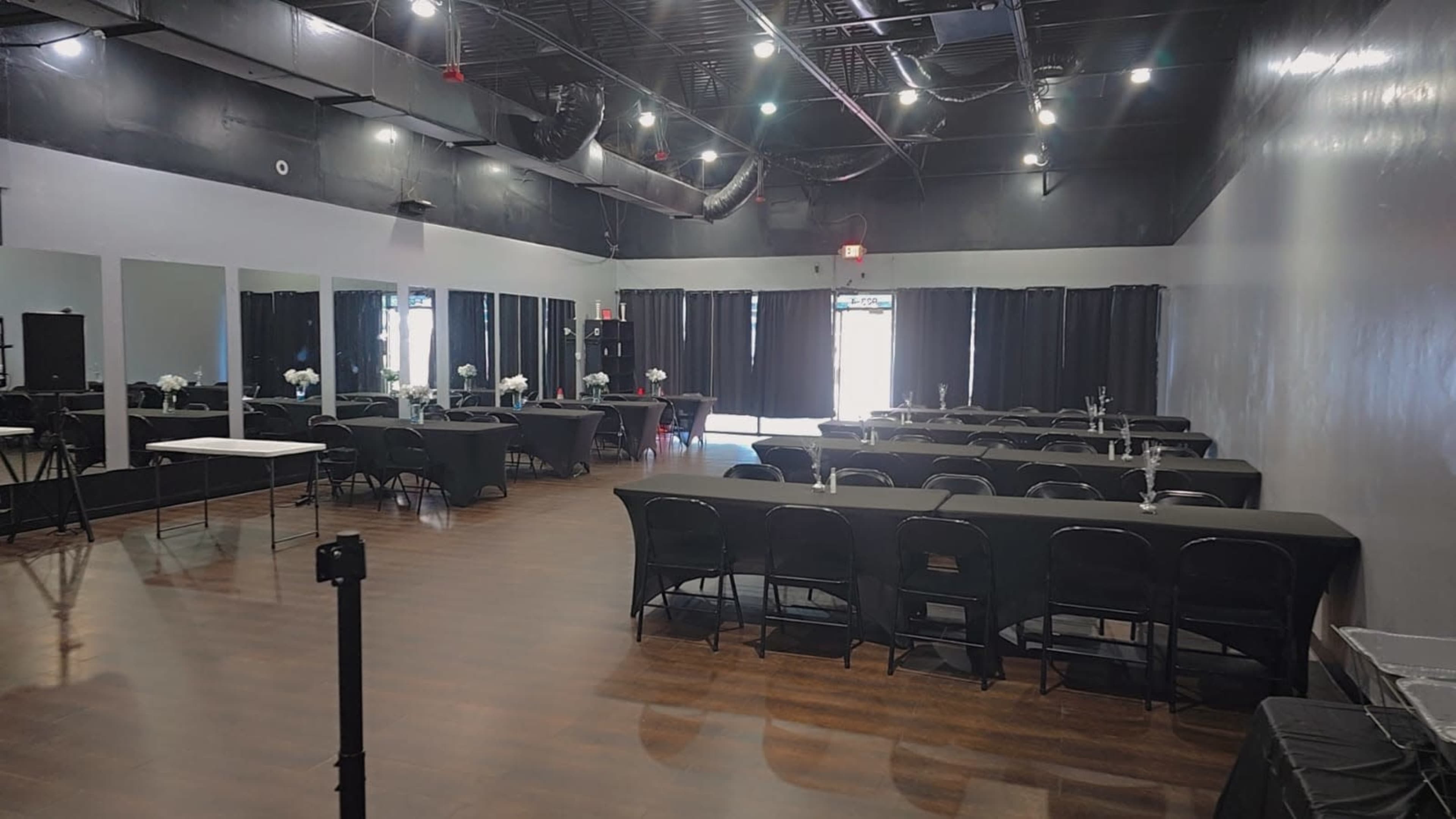 The image shows an empty event space with black tables and chairs arranged, mirrors on one wall, and black curtains covering the windows.