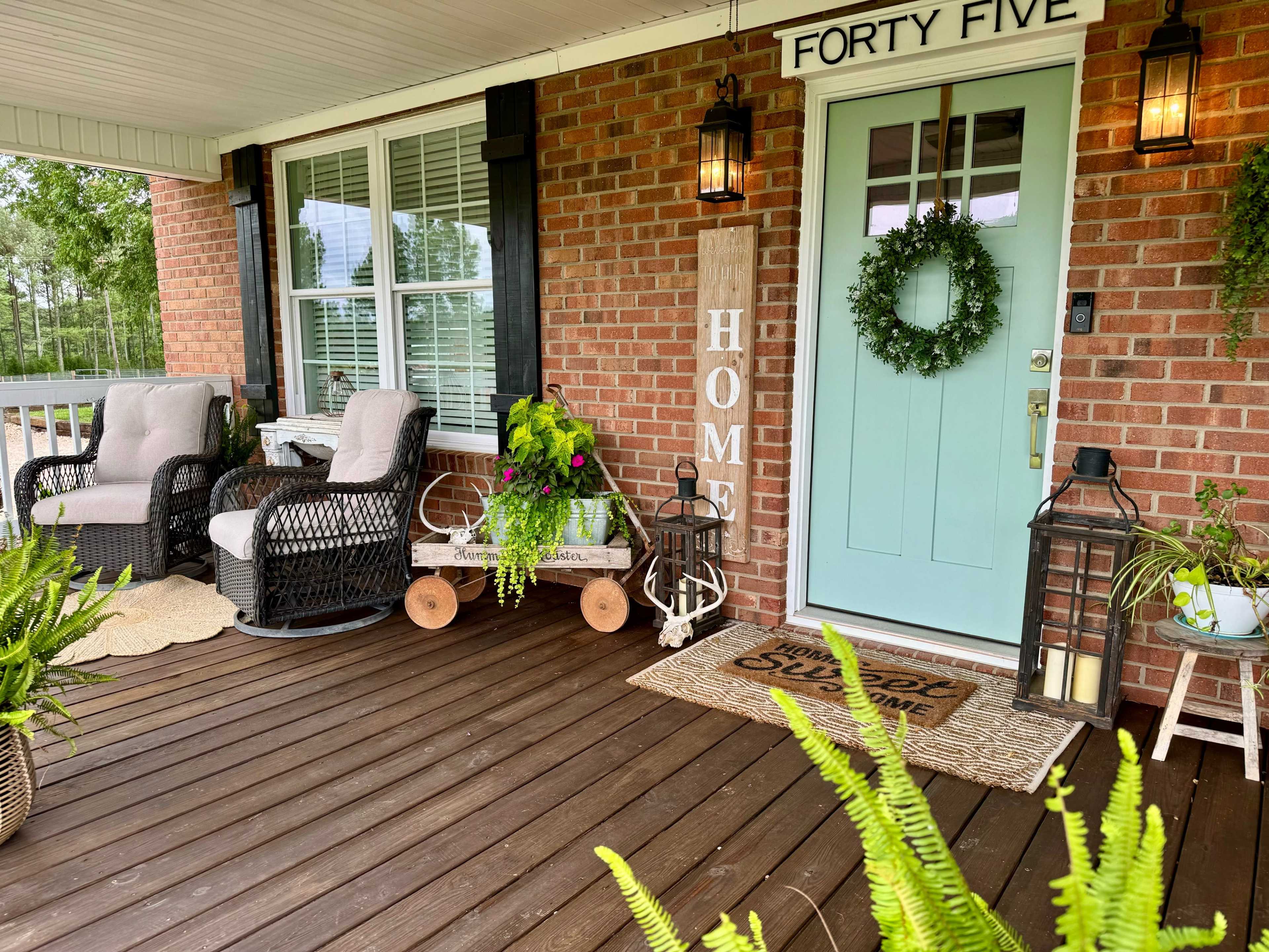 A front porch with a light blue door, potted plants, wicker chairs, and a welcome mat, surrounded by a brick facade.