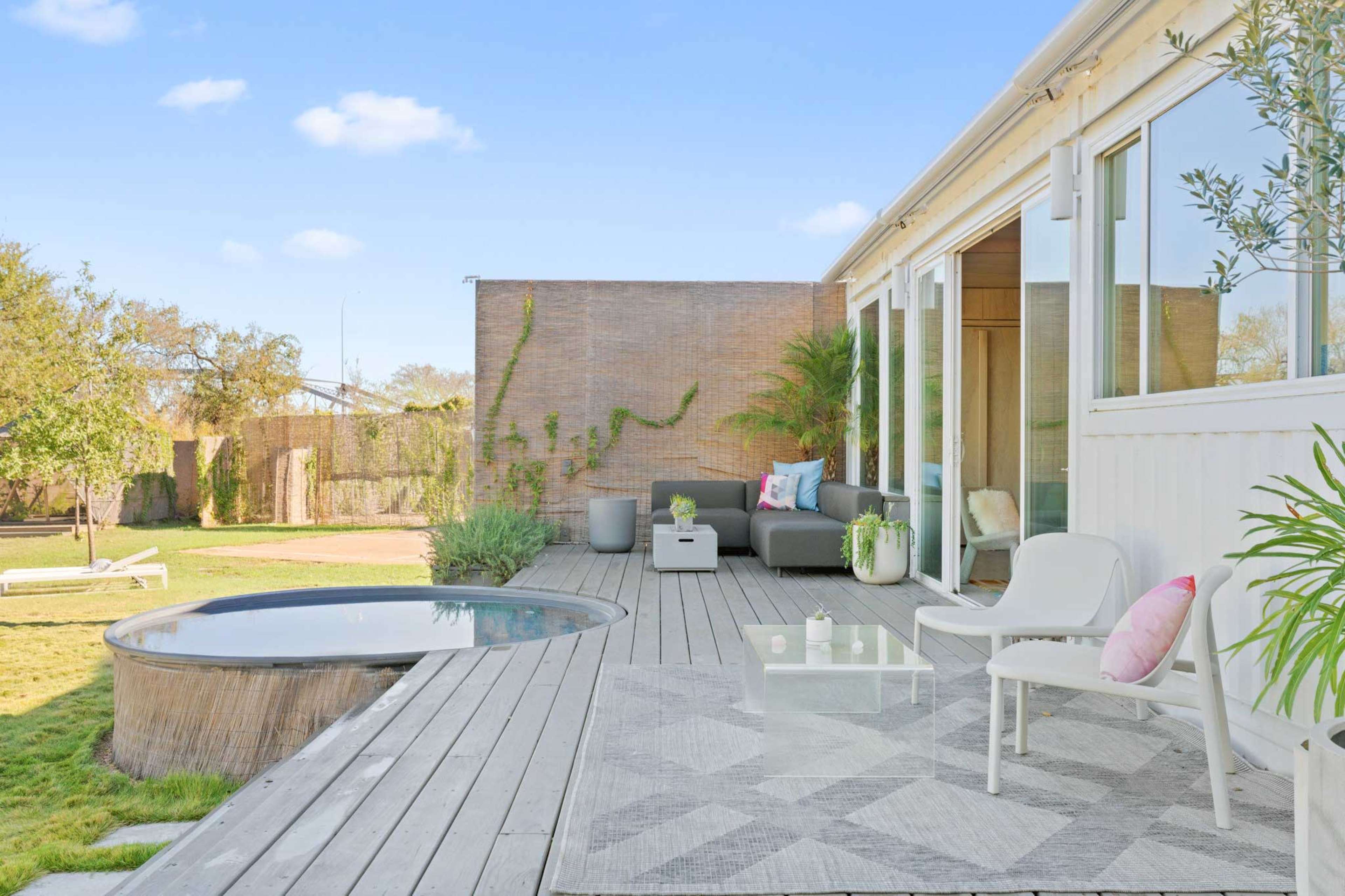 The image shows a modern outdoor deck featuring comfortable seating, a small round pool, and an arrangement of plants in a landscaped garden.