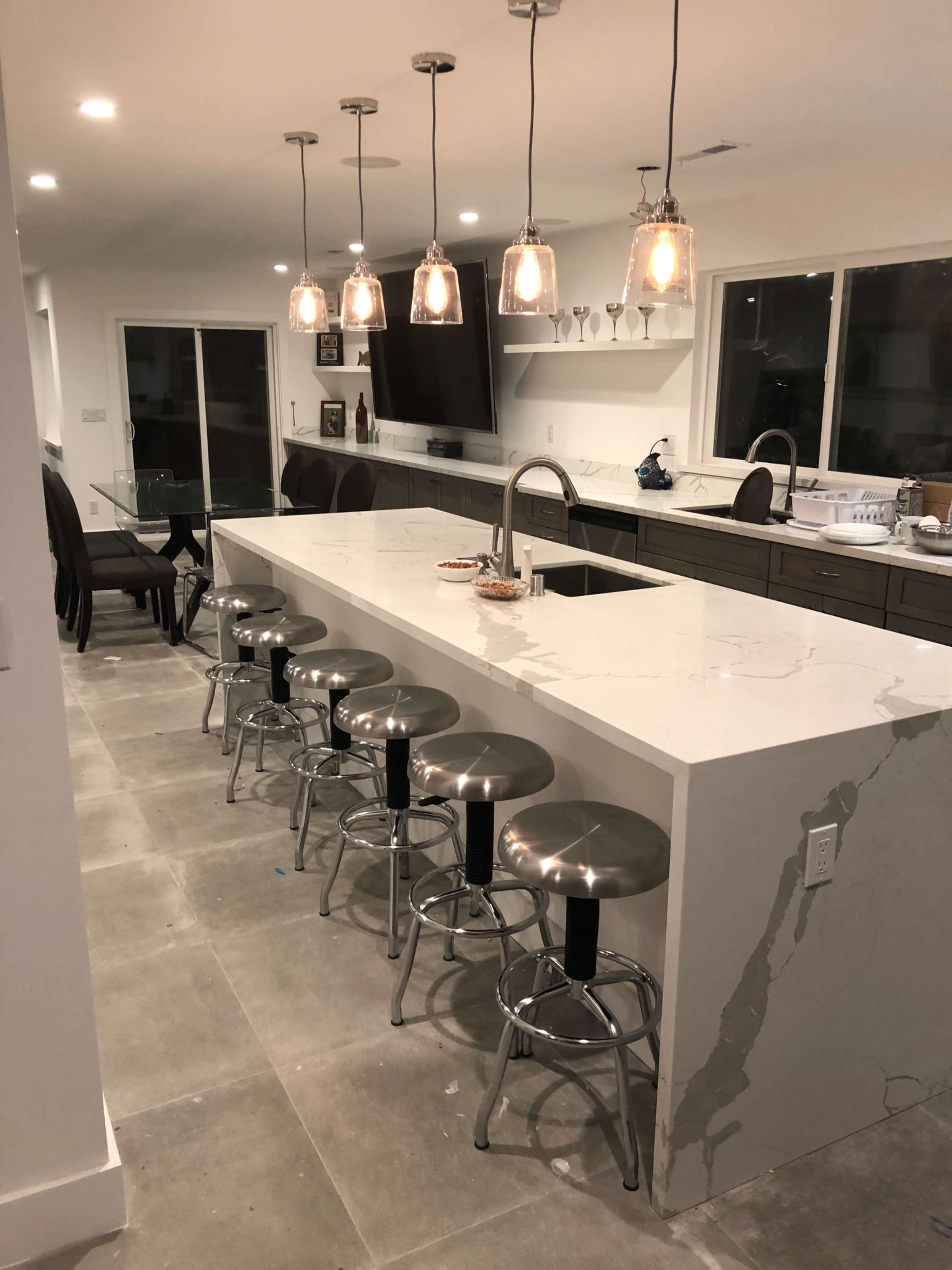 The image shows a modern kitchen with a long island featuring six metallic stools, illuminated by hanging pendant lights.