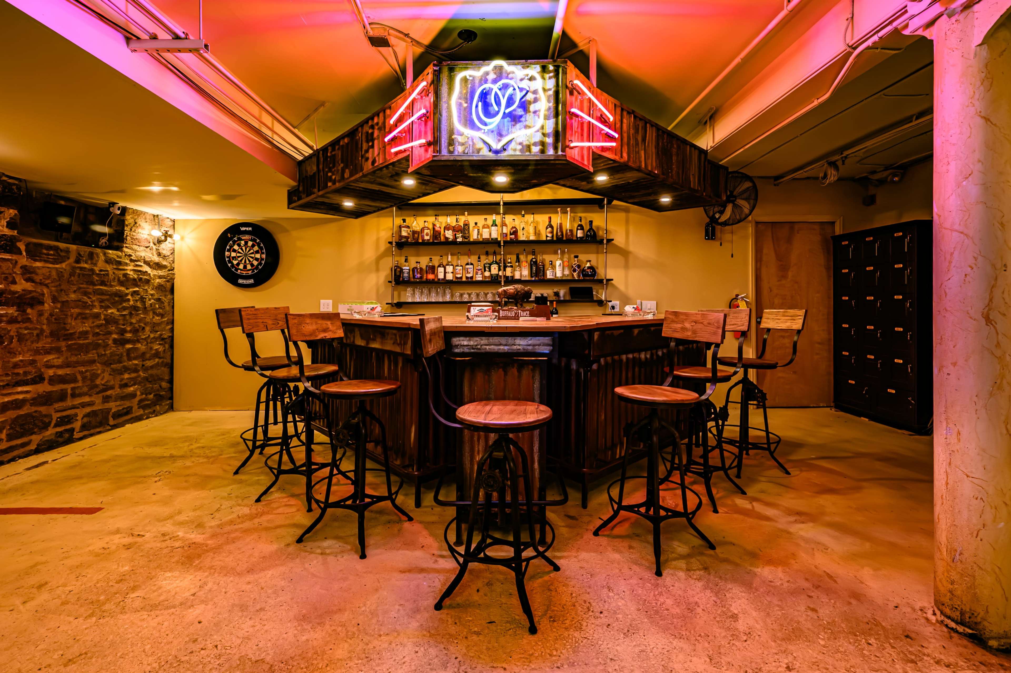 The image shows a dimly lit bar area featuring a wooden counter, several high stools, shelves stocked with bottles, and a neon sign overhead.