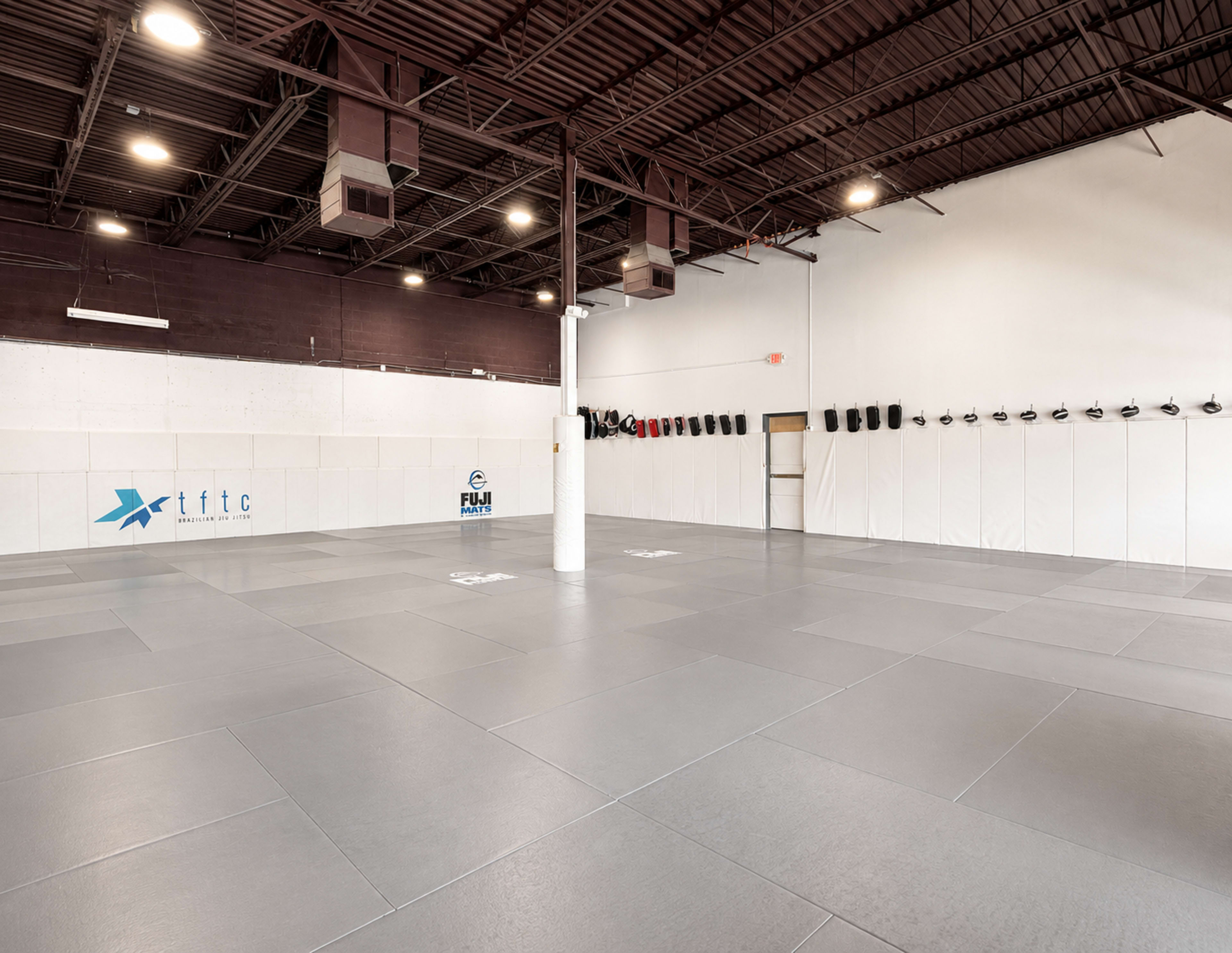 The image shows a spacious, empty martial arts training studio with gray flooring and padded walls, featuring some training equipment on the wall.