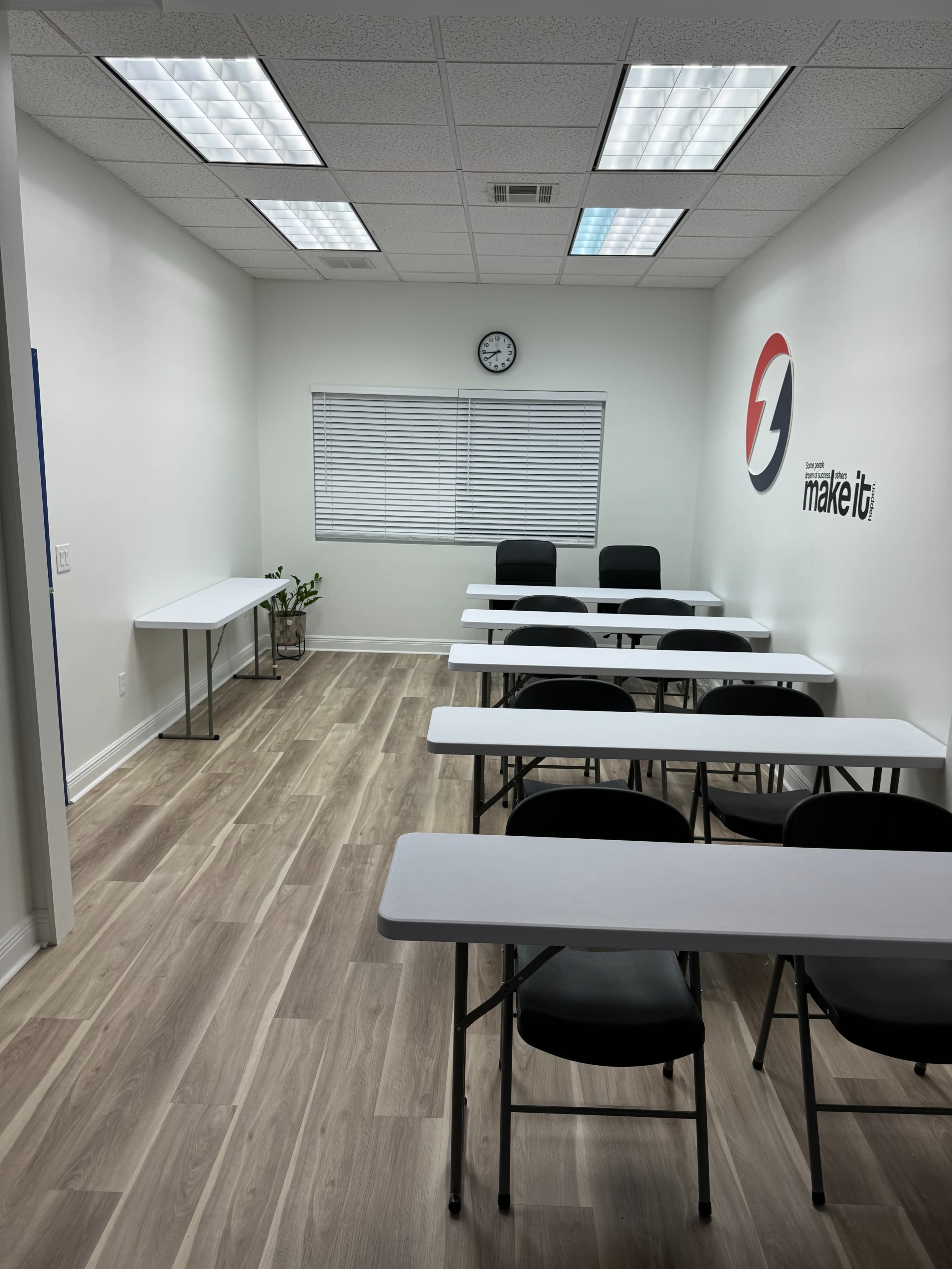 The image shows a classroom setup with several tables and chairs arranged neatly in a well-lit, minimalist space.