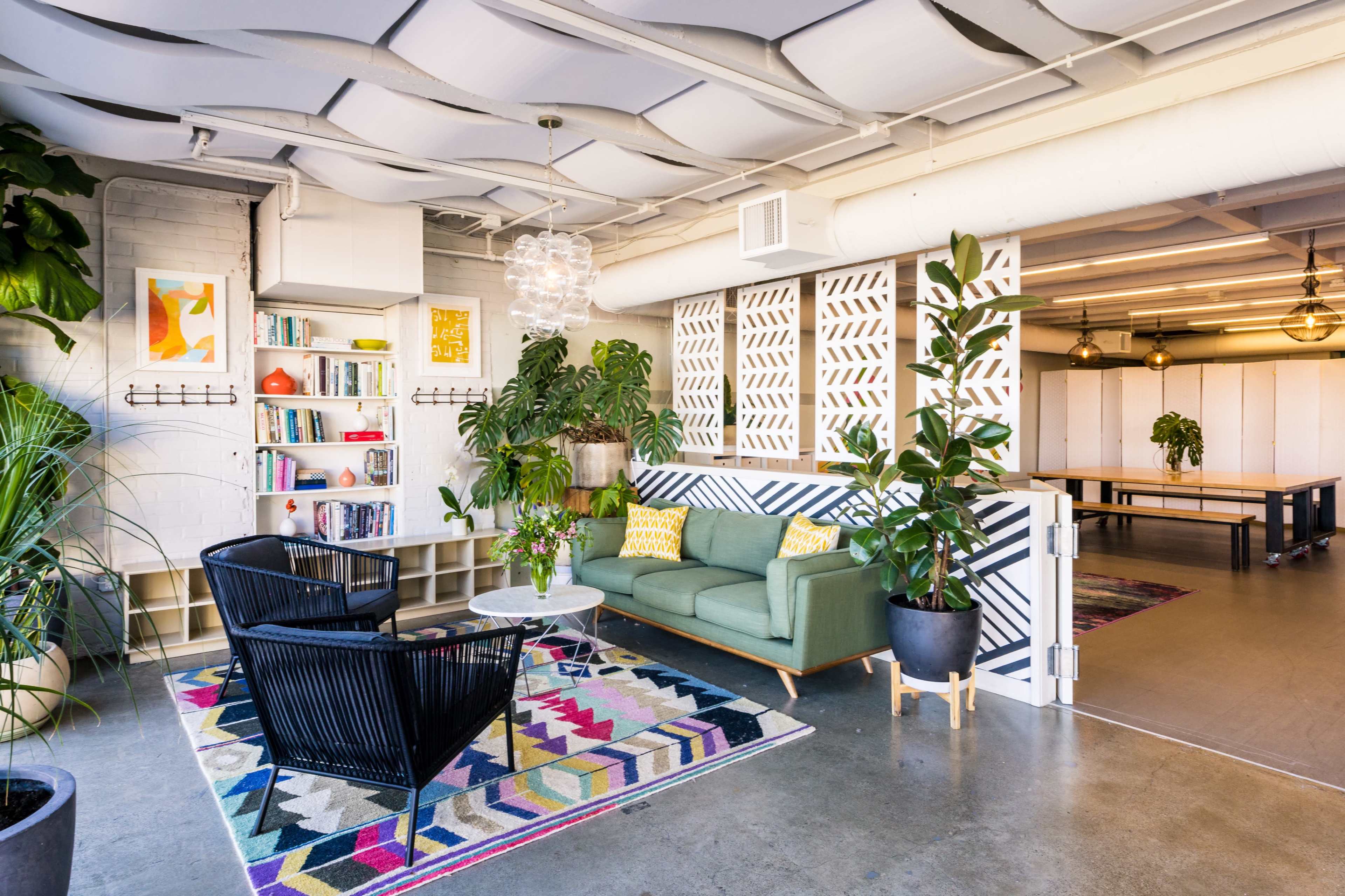 A modern office lounge area featuring a green couch, black chairs, a coffee table, a bookshelf filled with books, and various plants against a backdrop of patterned walls and overhead lighting.