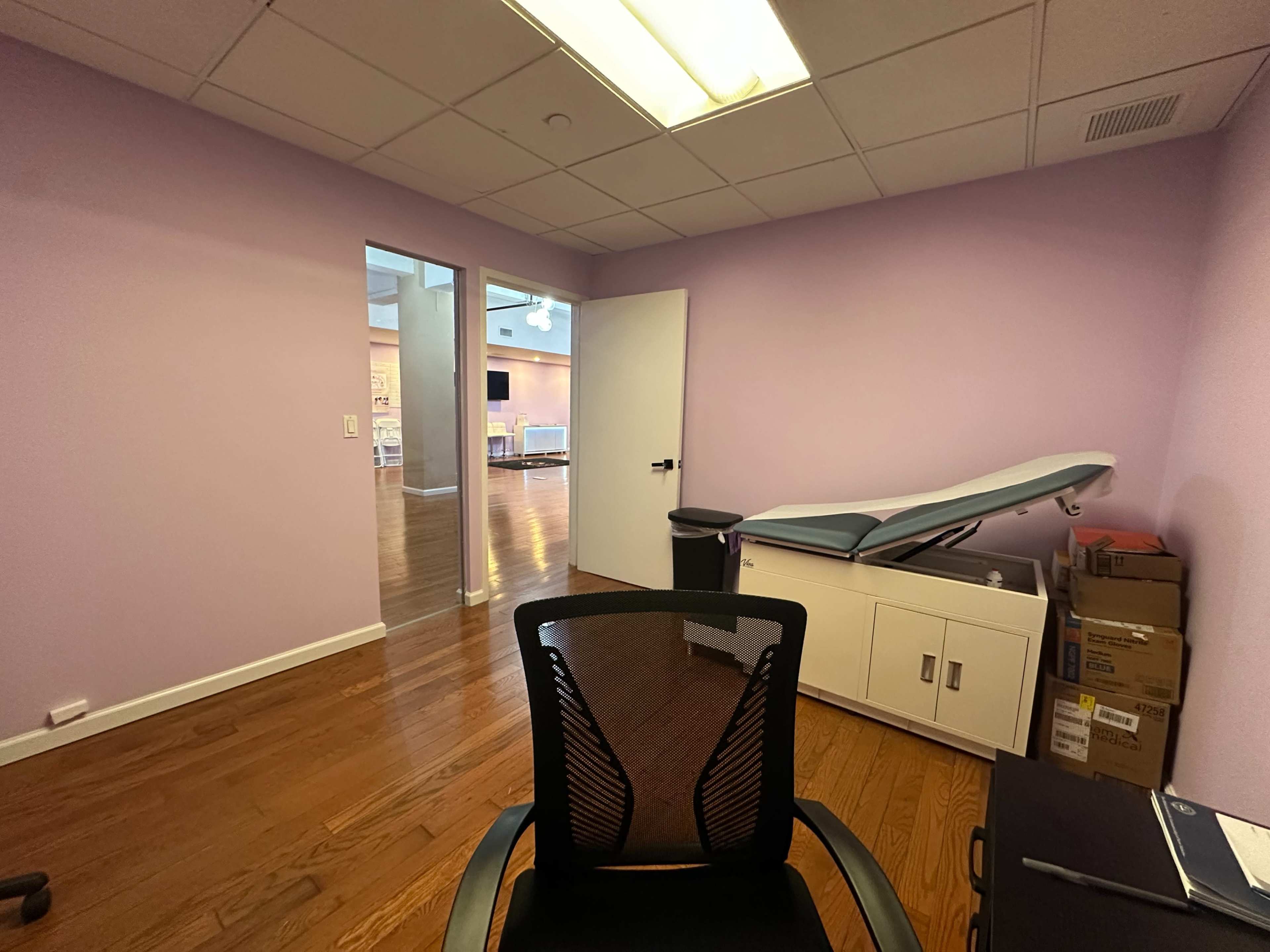 The image shows a room with purple walls, hardwood flooring, a chair facing a desk, a medical examination table, and a doorway leading to another space.