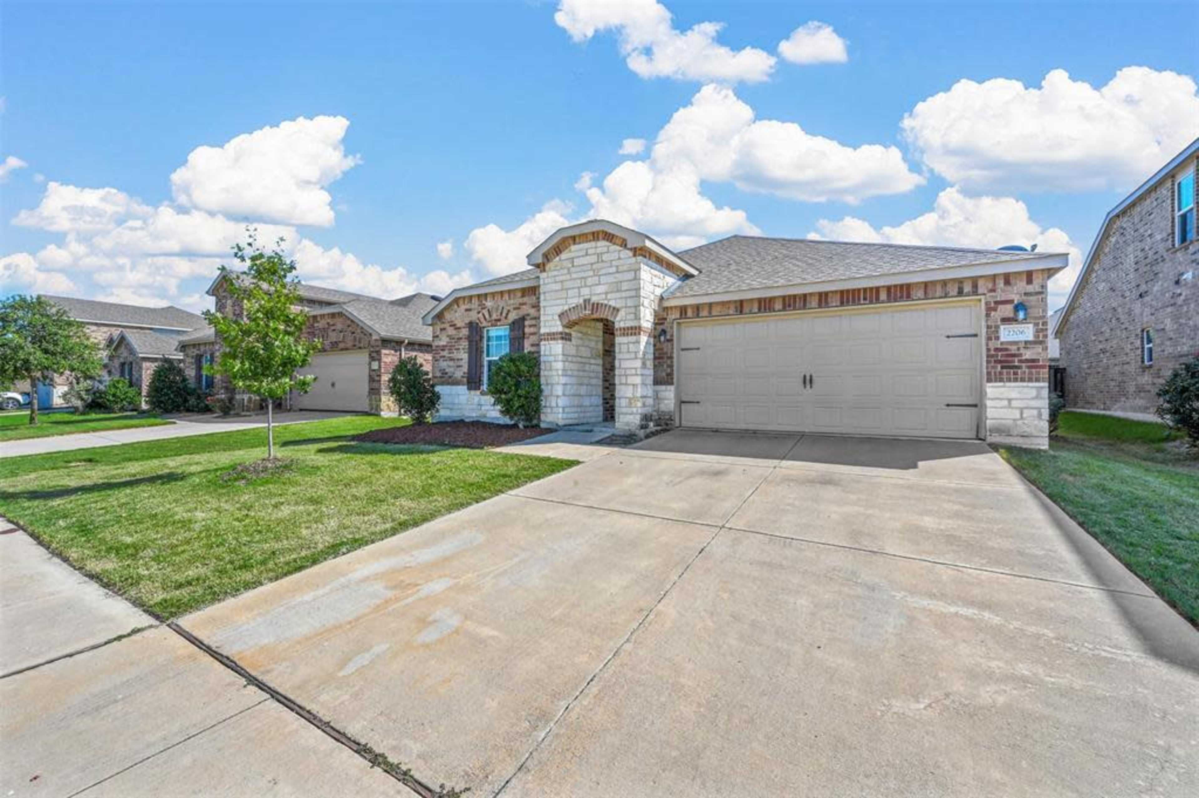 The image shows a suburban home with a stone and brick exterior, two-car garage, and a concrete driveway surrounded by green grass and a few trees.