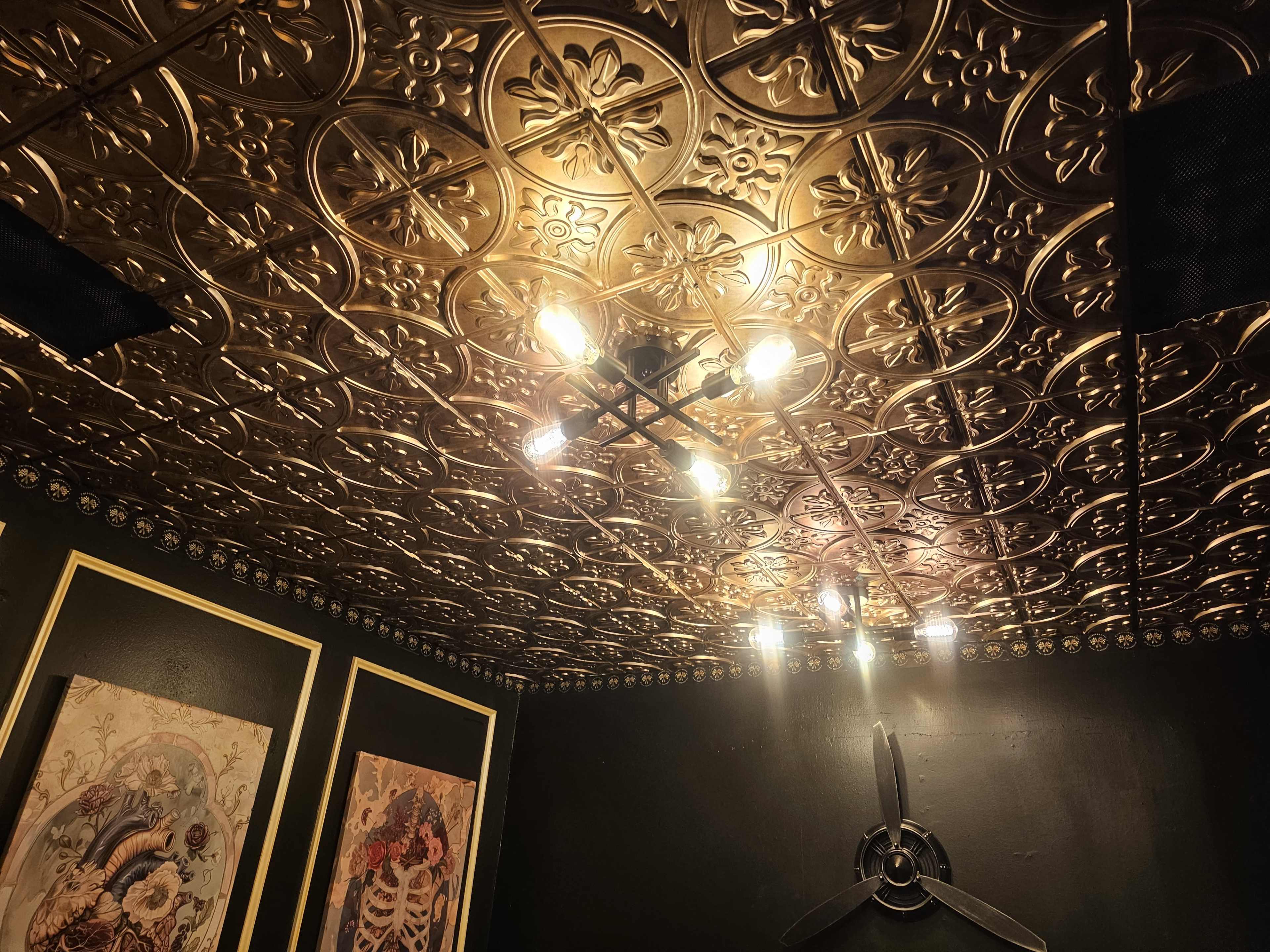 The image shows a ceiling with ornate gold detailing, illuminated by multiple light fixtures, alongside two framed artworks on dark walls.