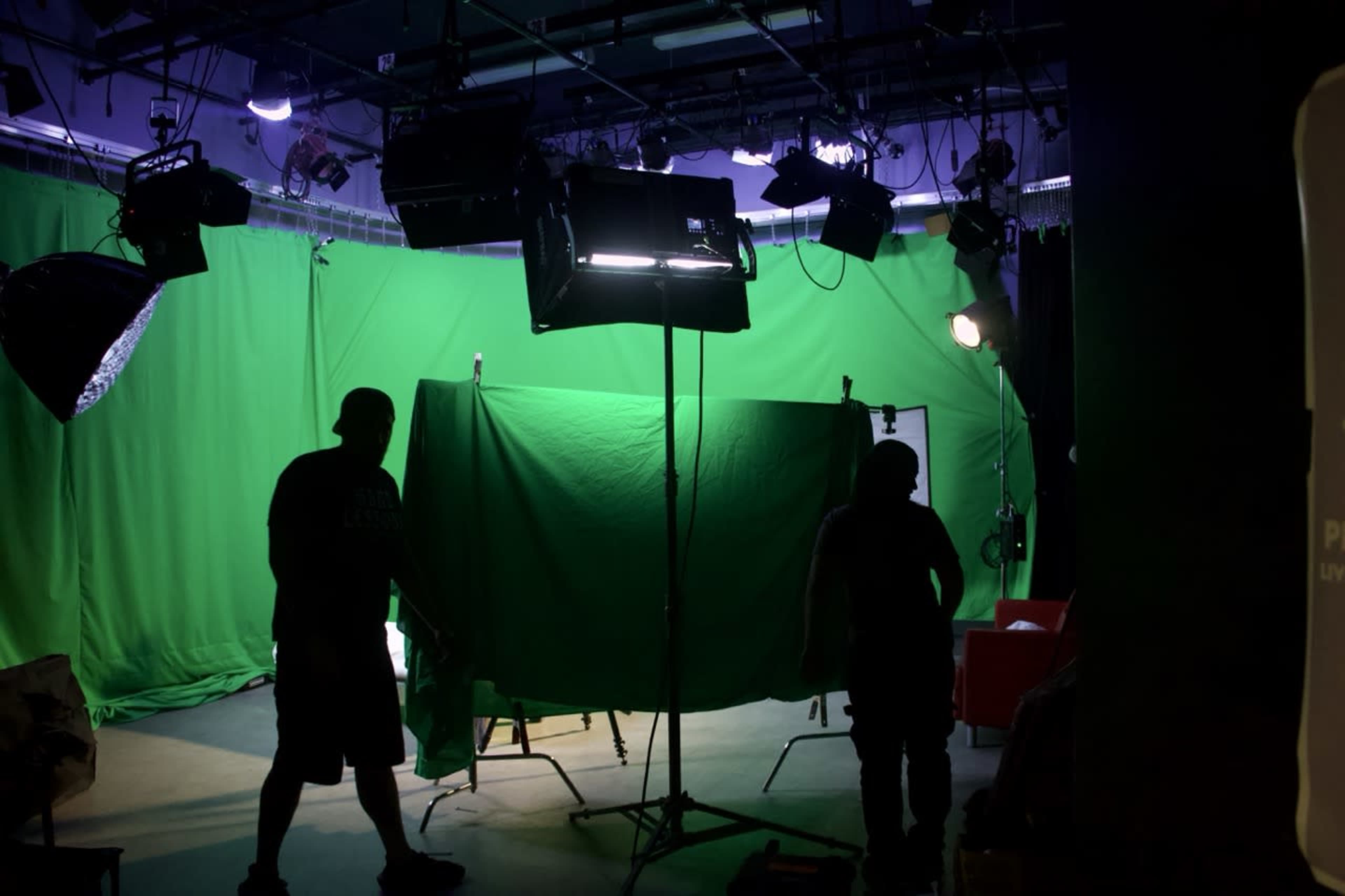 A film crew sets up lighting and green screens in a studio for video production.