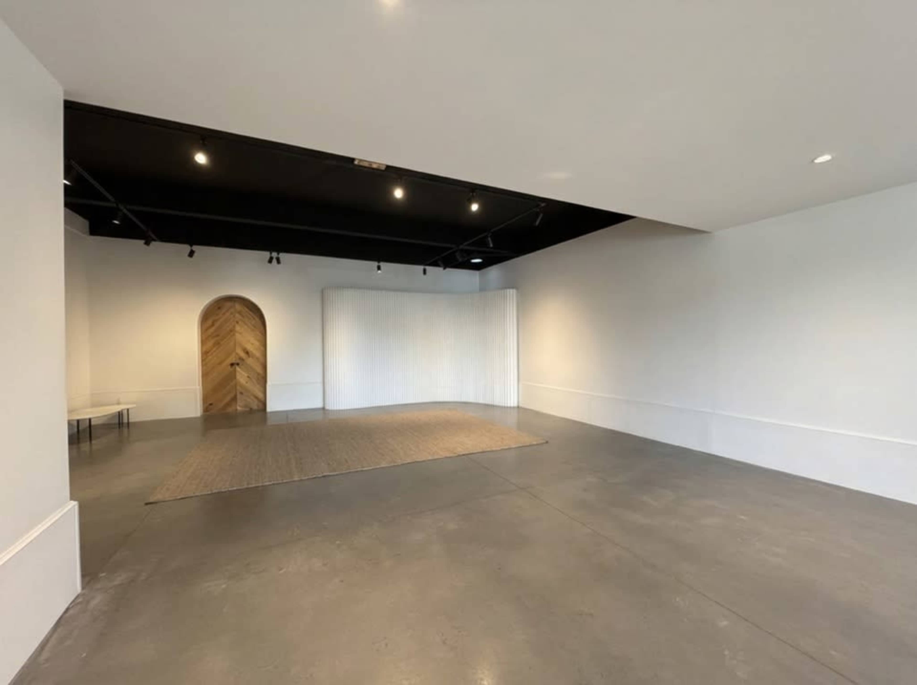 The image shows a spacious, minimalist interior with concrete flooring, a textured wooden archway, and a soft fabric partition.