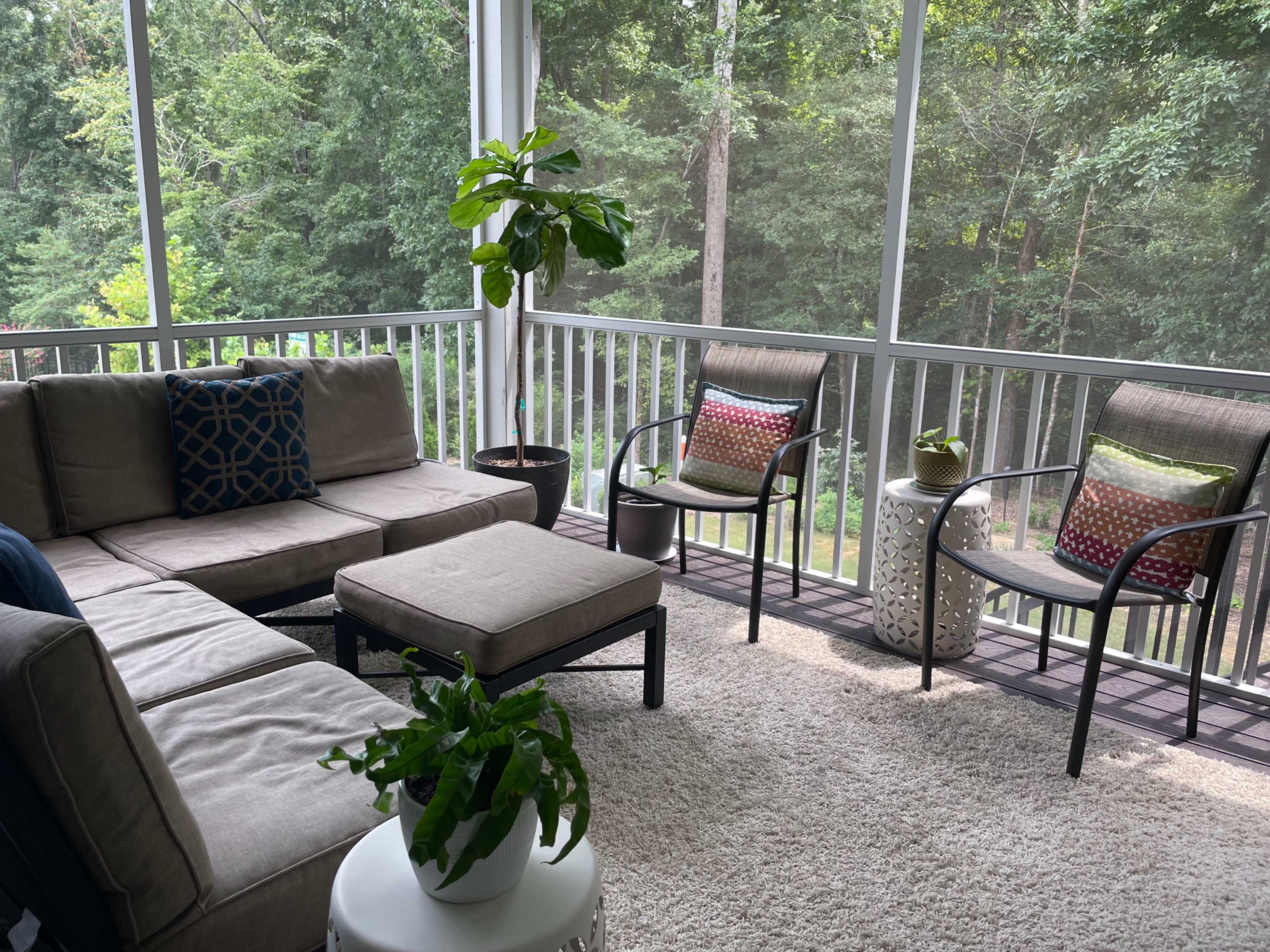 A screened-in porch features a sectional sofa, two chairs with colorful cushions, a coffee table, and potted plants, set against a backdrop of dense greenery.