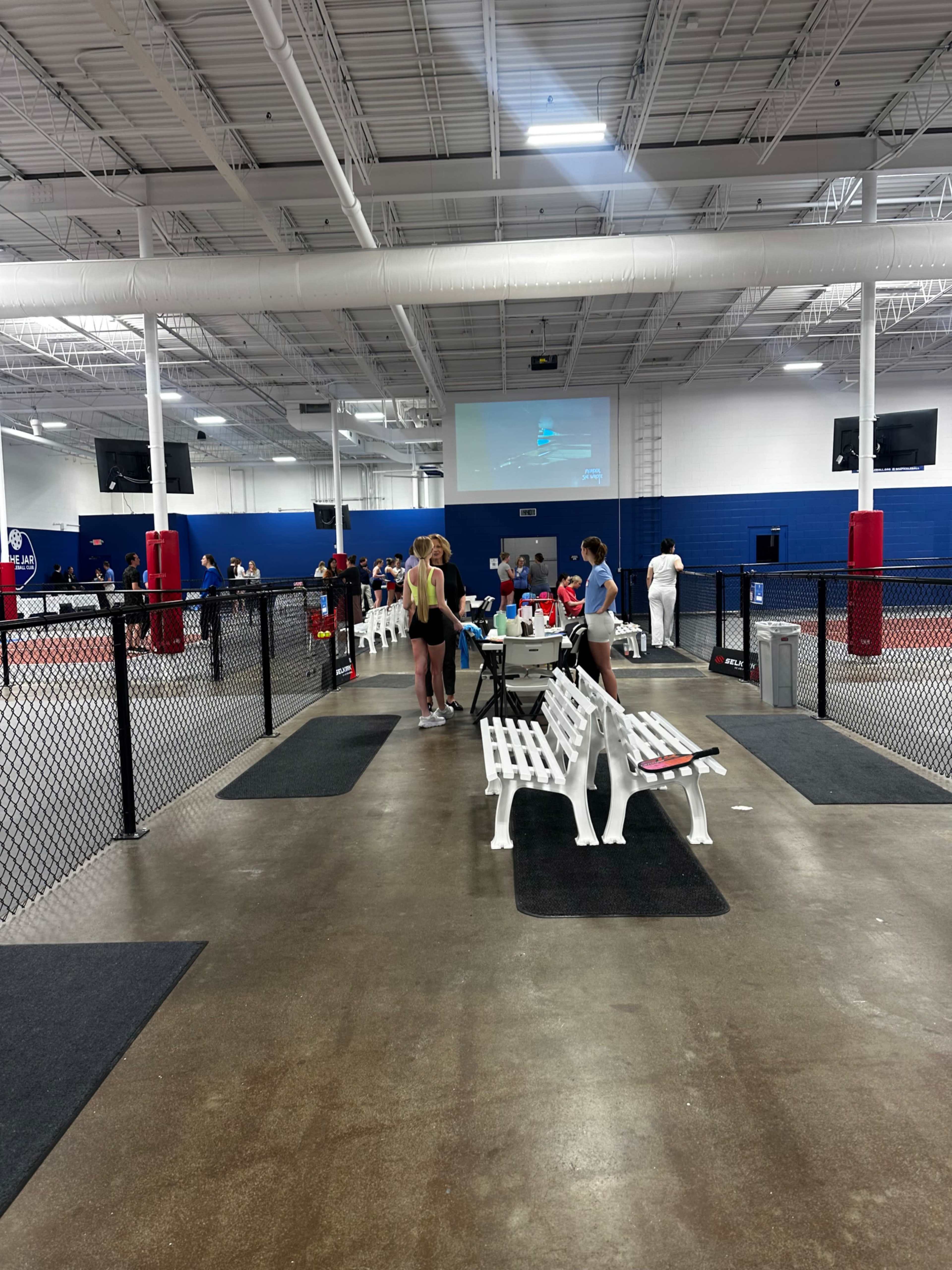 The image shows an indoor sports facility with people engaging in various activities, benches along the sidelines, and a large screen displaying content in the background.