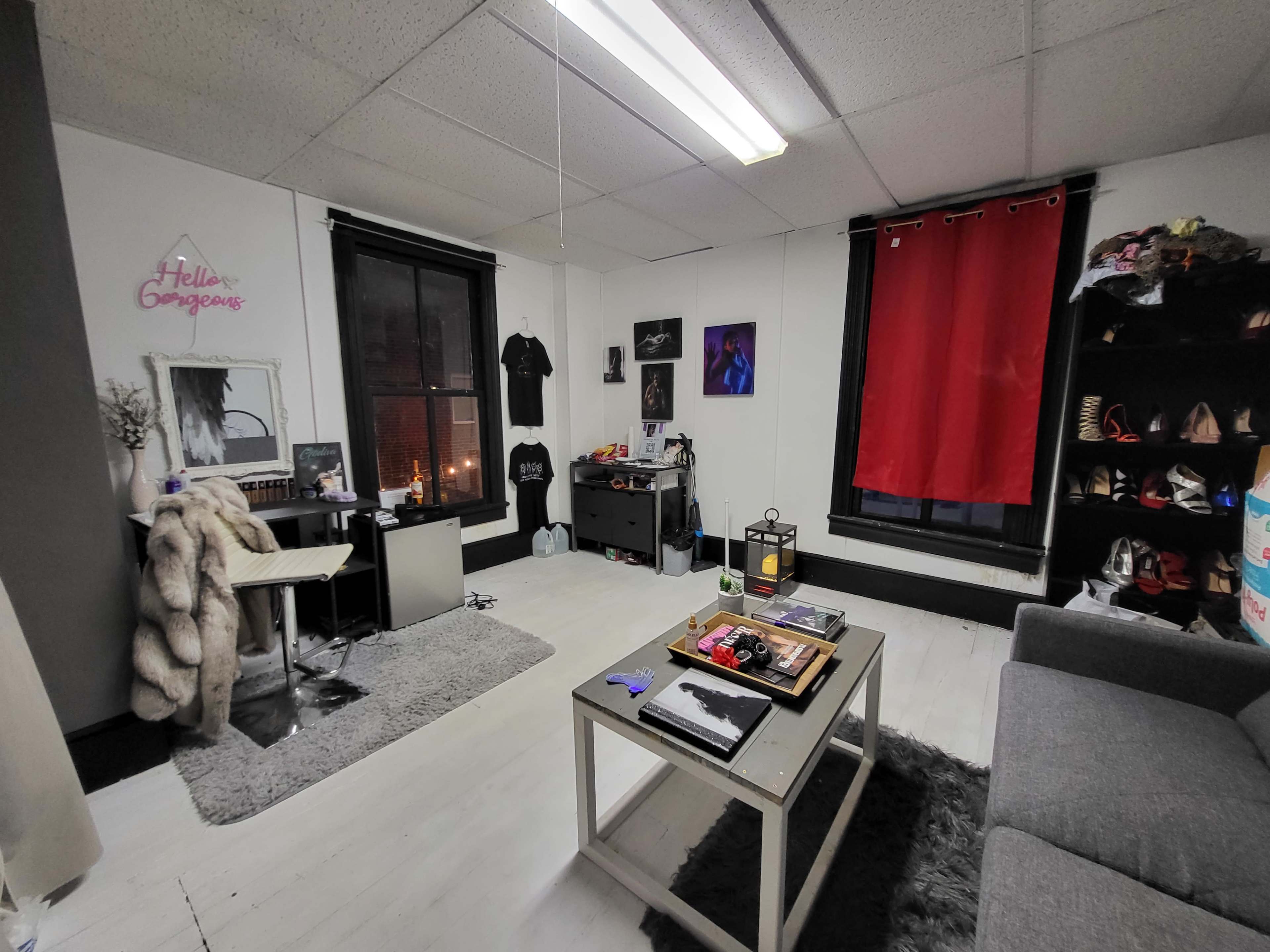 The image shows a small, cluttered room featuring a gray rug, a coffee table, a red curtain, and a shelf filled with shoes.