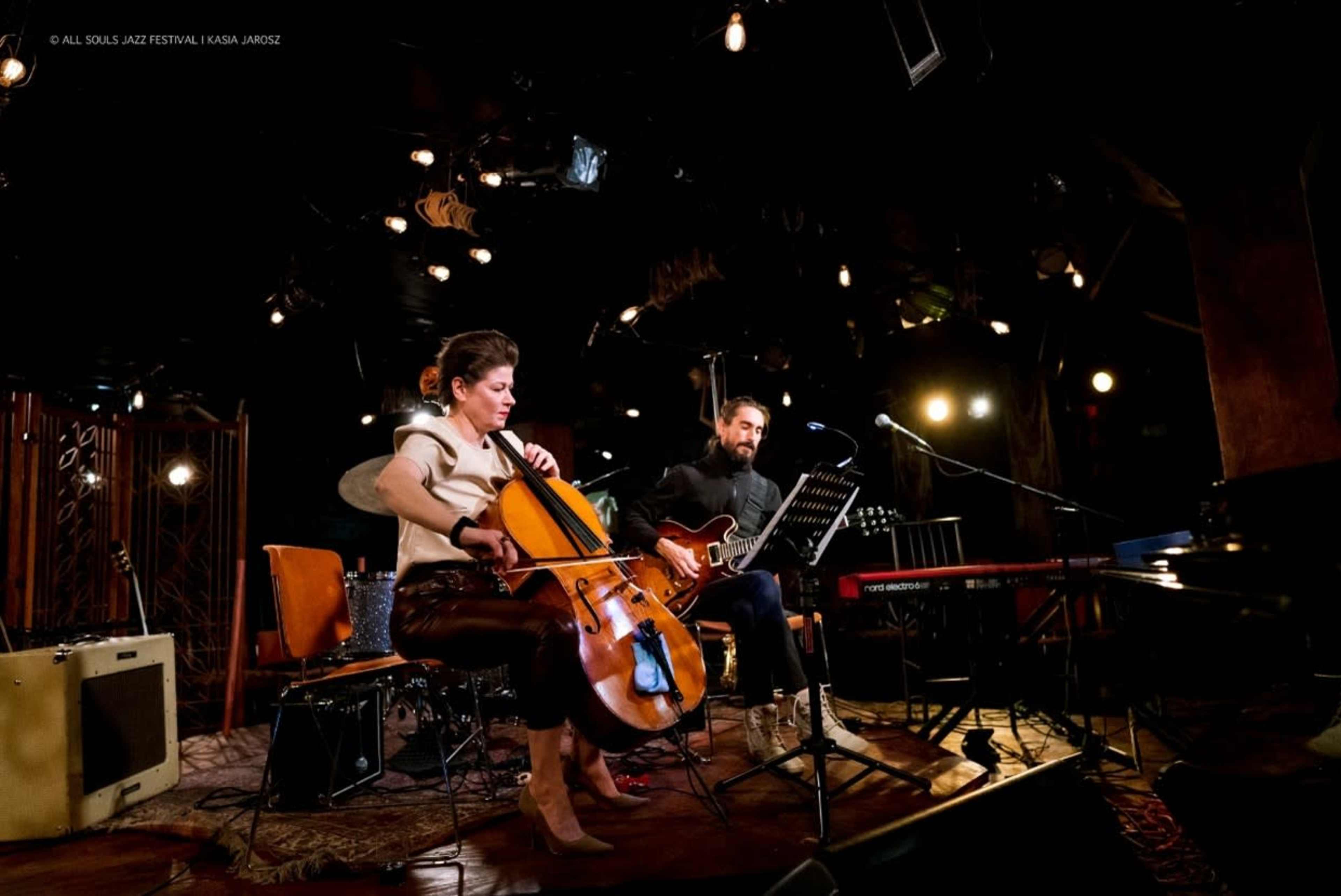 Two musicians perform on stage, one playing a cello while the other plays an electric guitar, surrounded by various instruments and stage lighting.