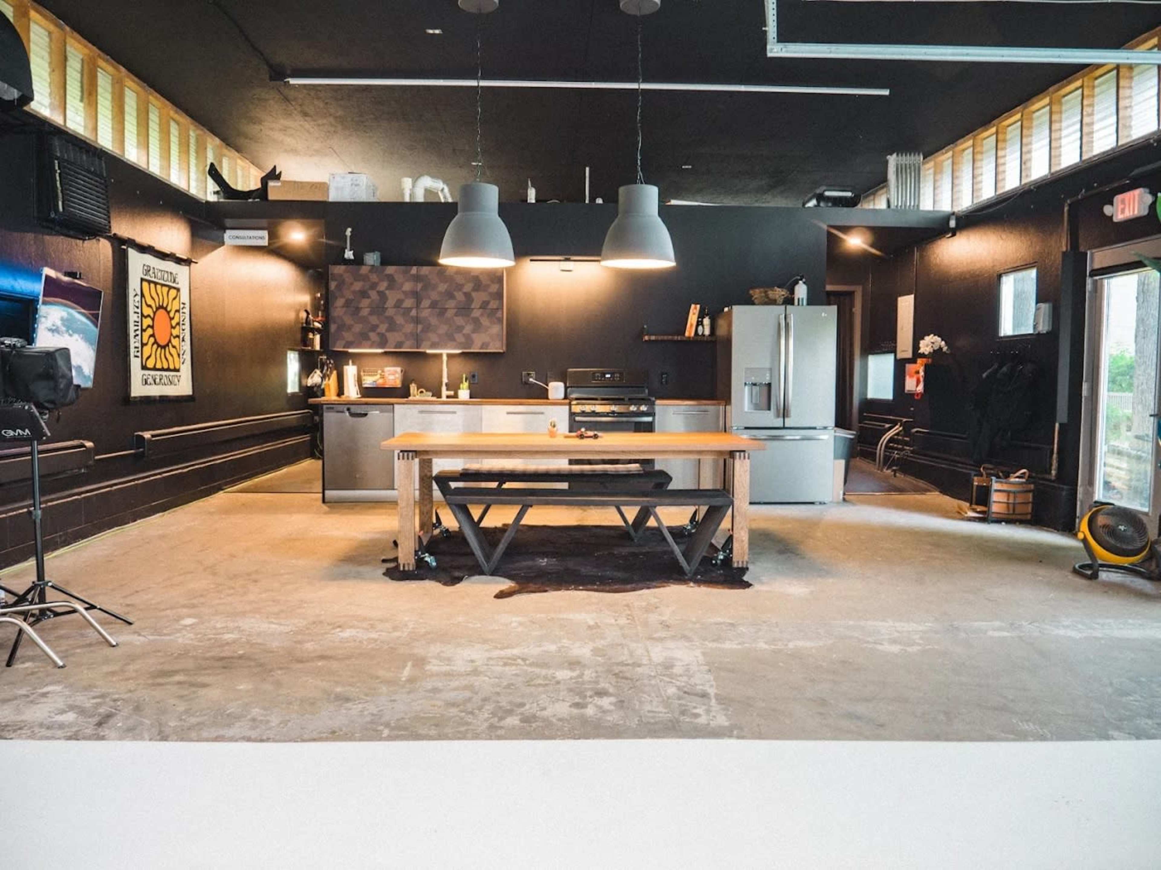 The image shows a modern kitchen with a large wooden table in the center, stainless steel appliances, and dark walls illuminated by pendant lighting.
