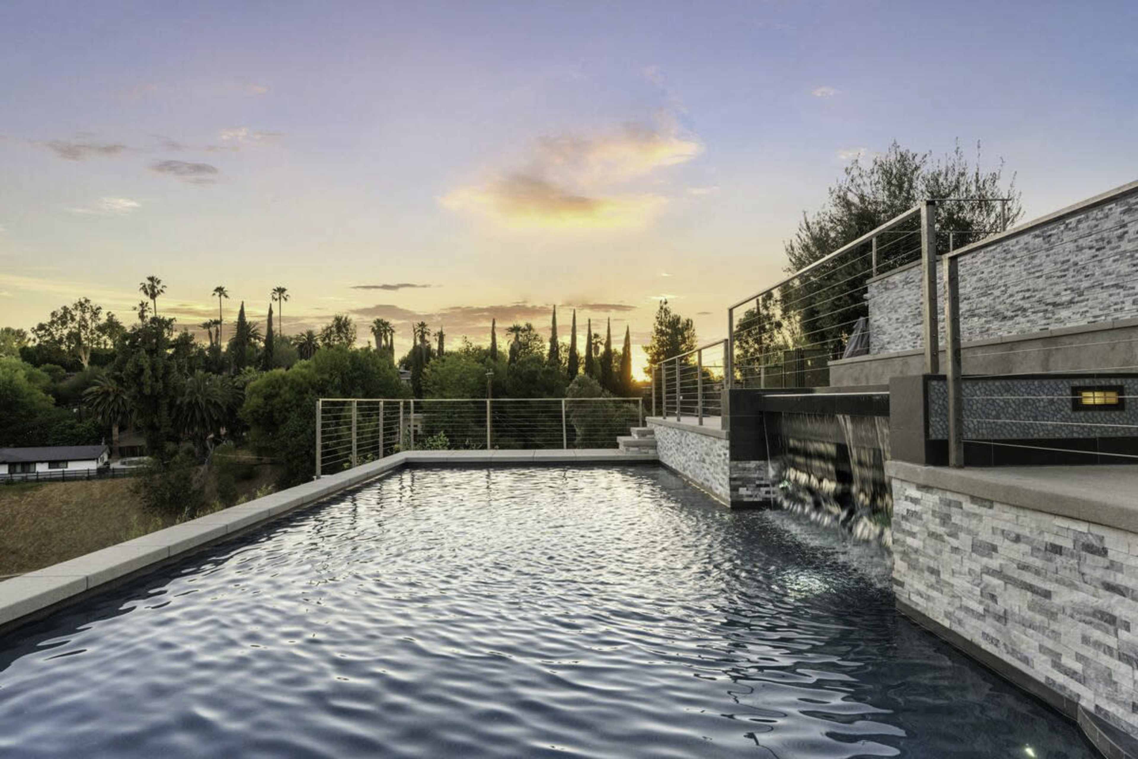 The image shows a modern infinity pool with a stone terrace overlooking a lush landscape at sunset.