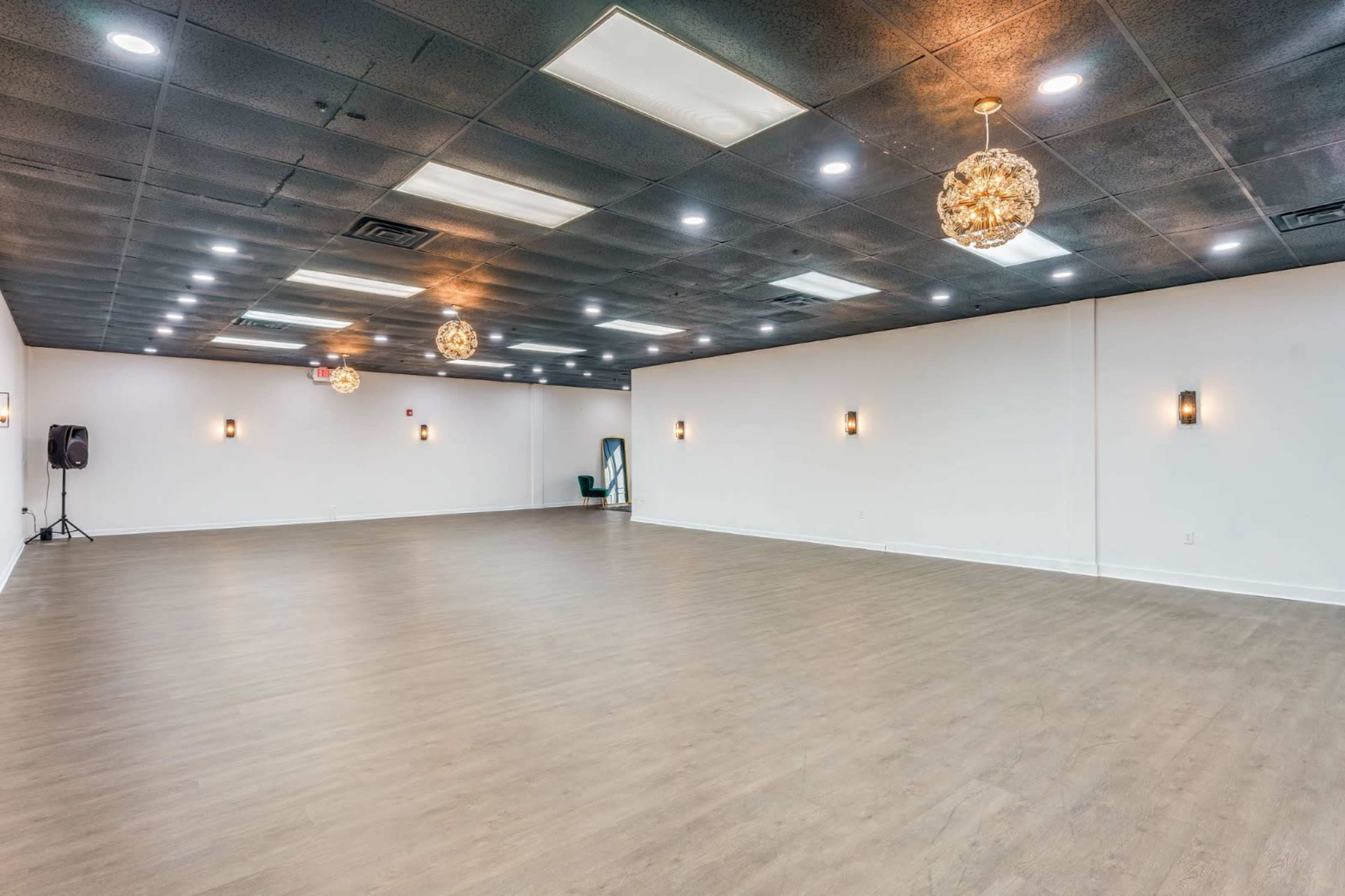 The image shows a spacious, empty room with a wooden floor, modern lighting fixtures on the ceiling, and simple wall sconces.