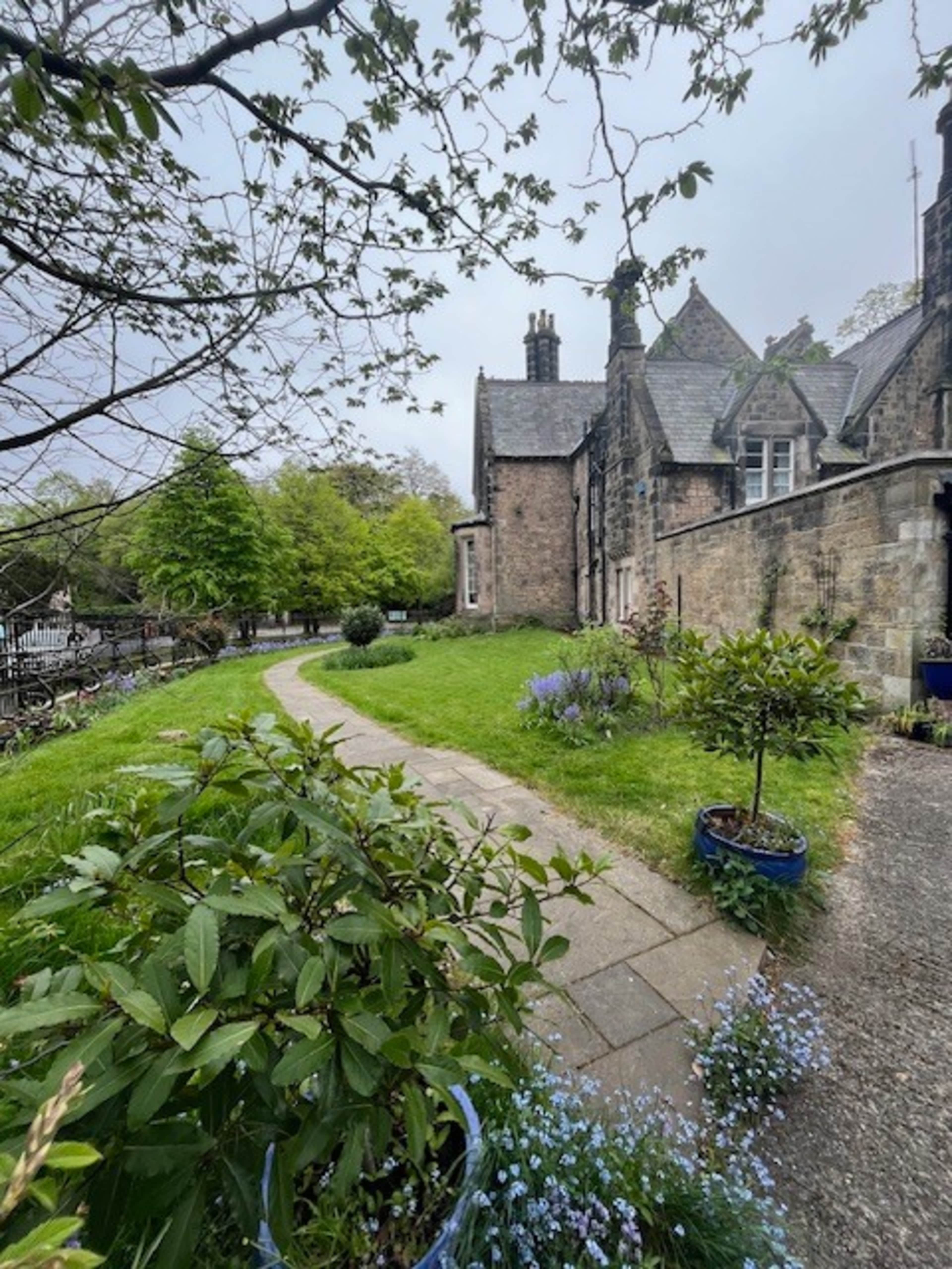The image shows a pathway lined with greenery leading to a historic stone building with multiple chimneys and windows.