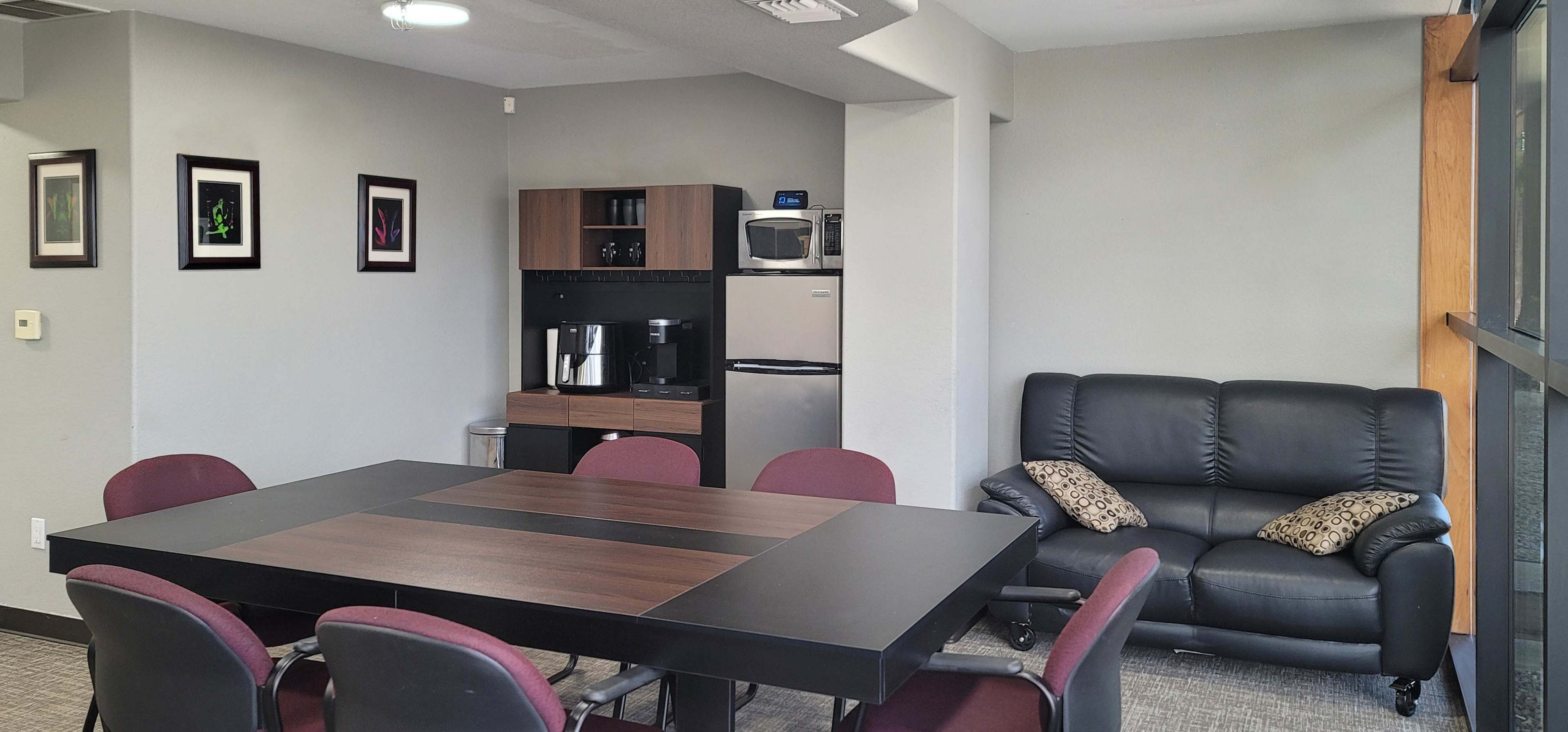 The image depicts a multi-purpose room with a large table surrounded by chairs, a kitchenette in the background, and a seating area with a couch.