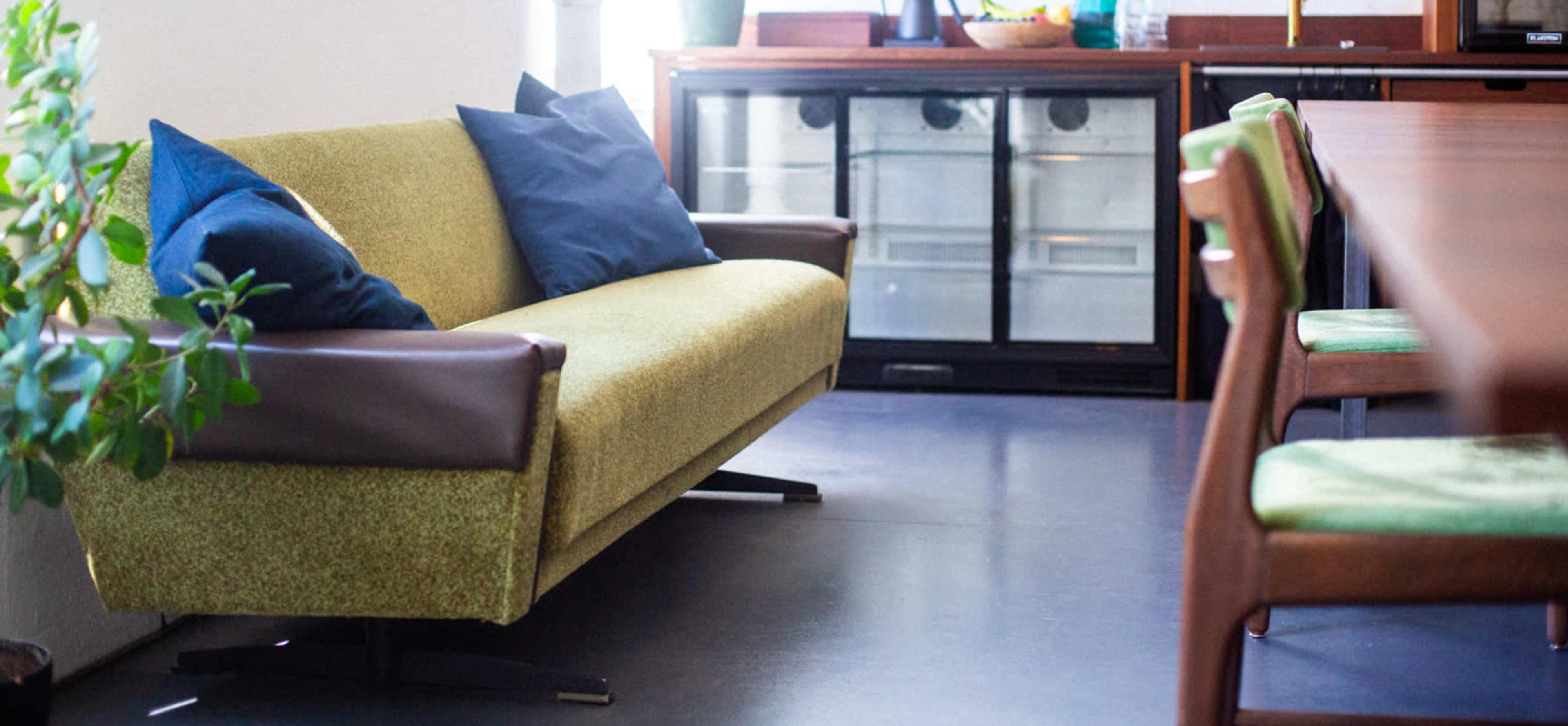 A green couch with blue pillows sits next to a dining table and a large refrigerator in a well-lit room.