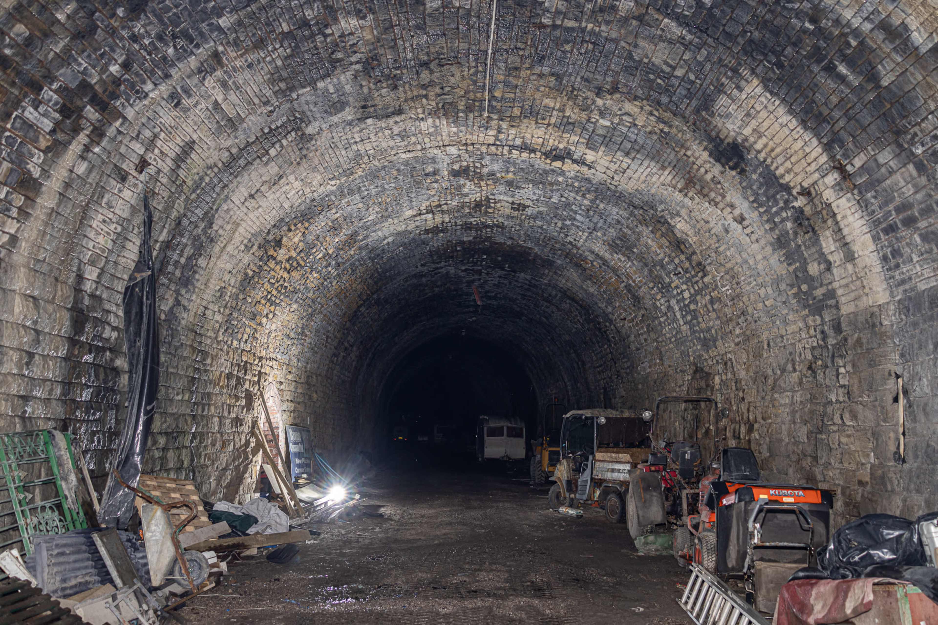 An arched stone tunnel interior features various pieces of machinery and equipment along one side, illuminated by artificial light.