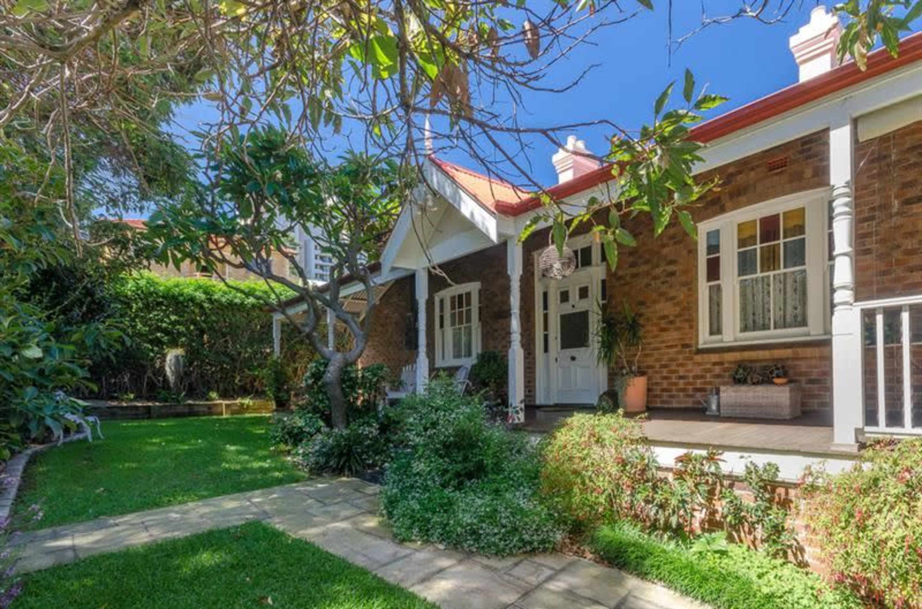 A brick house with a front porch and garden, featuring a pathway, trees, and lush greenery.