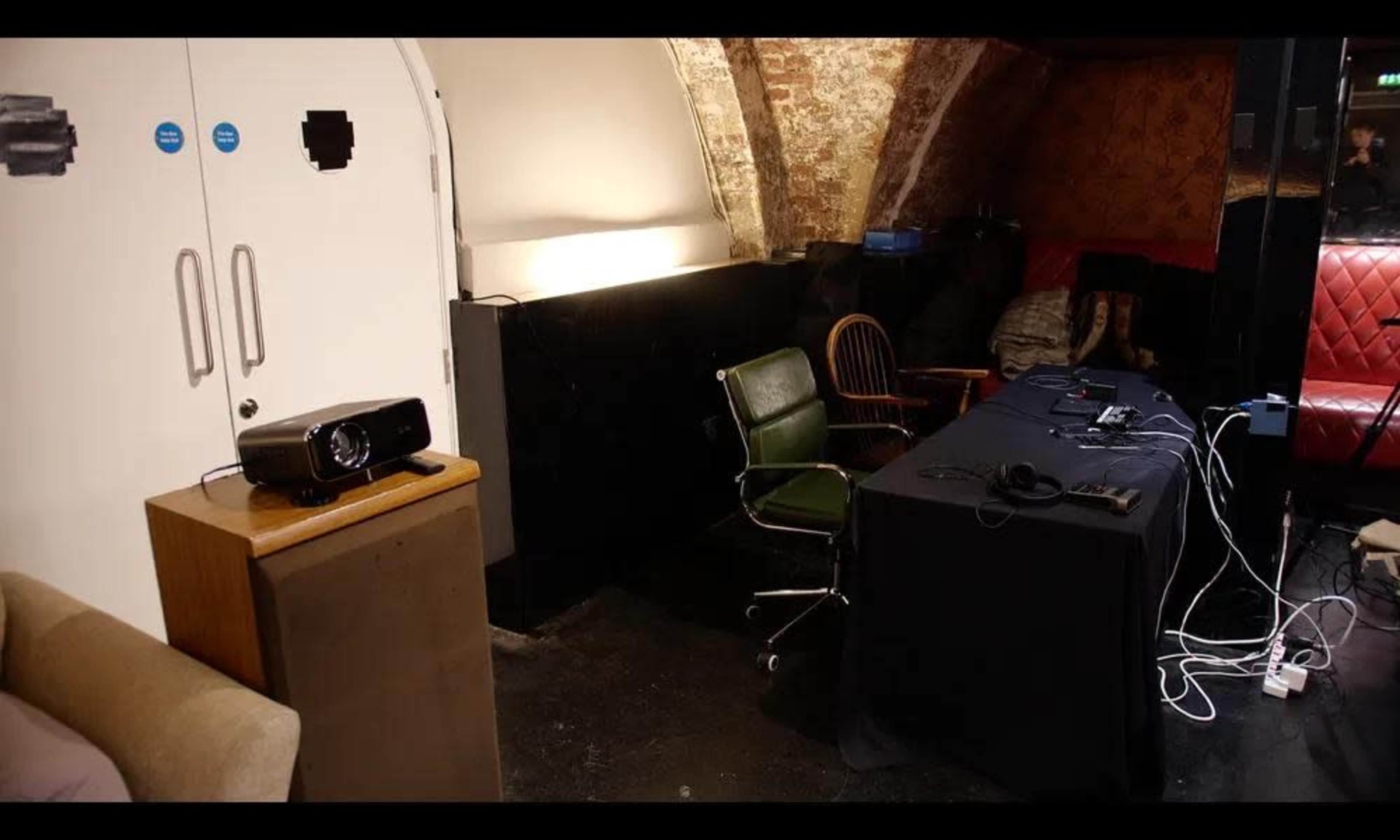 The image shows a dimly lit room with exposed brick walls, featuring a projector on a small table next to a green chair and a black table cluttered with cables and equipment.