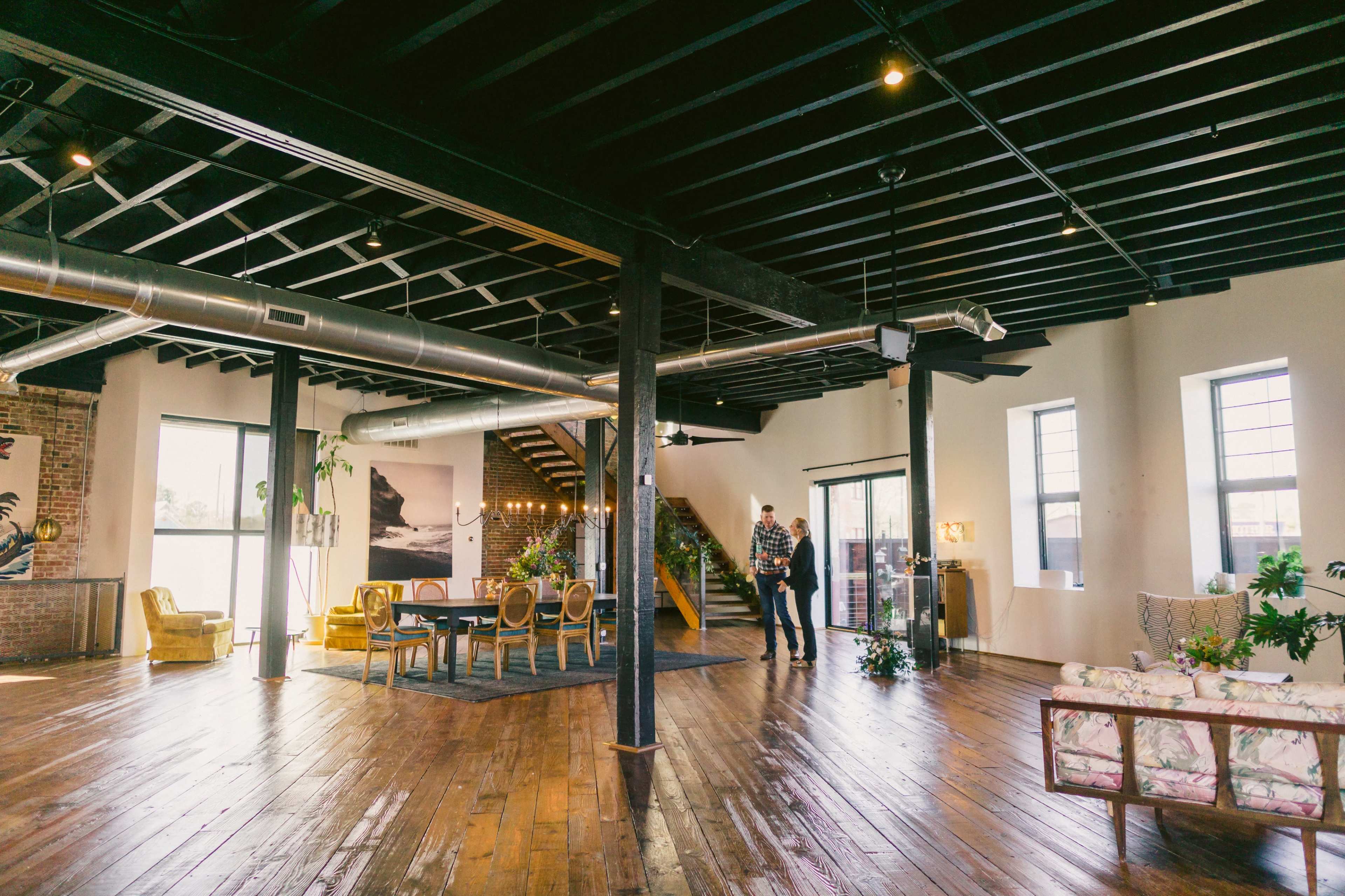 A spacious modern interior with wooden floors, large windows, a dining area, and a staircase leading to an upper level.